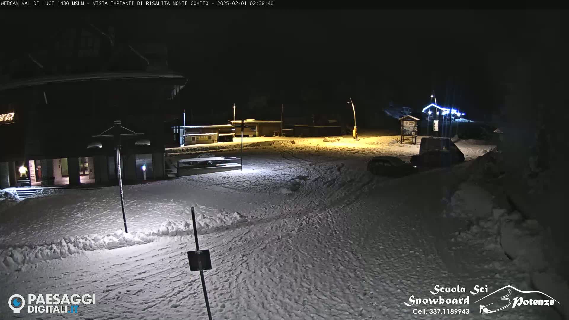 A snow-covered nighttime scene shows a building complex illuminated by artificial light, with a few vehicles parked nearby.