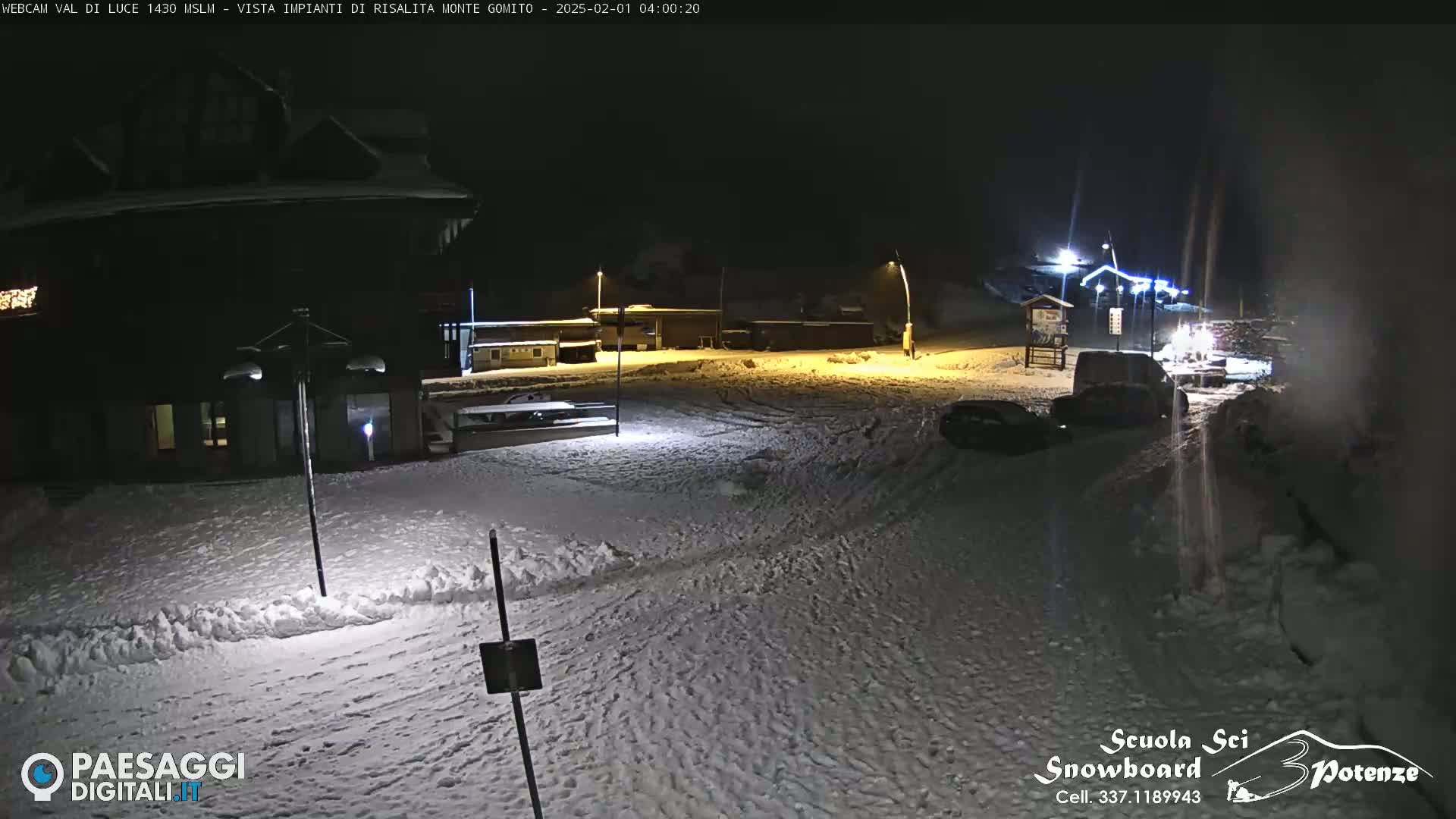 A snow-covered parking lot at night, illuminated by artificial lights, with several vehicles parked and buildings in the background.