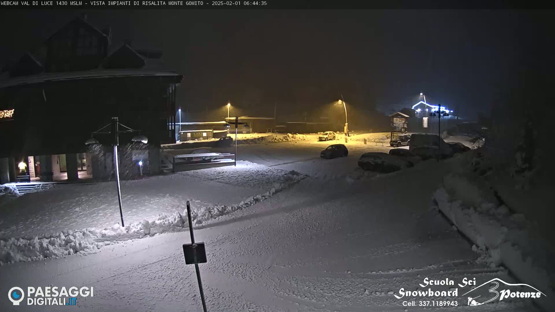 A snow-covered parking lot and building are illuminated at night by streetlights during a light snowfall.