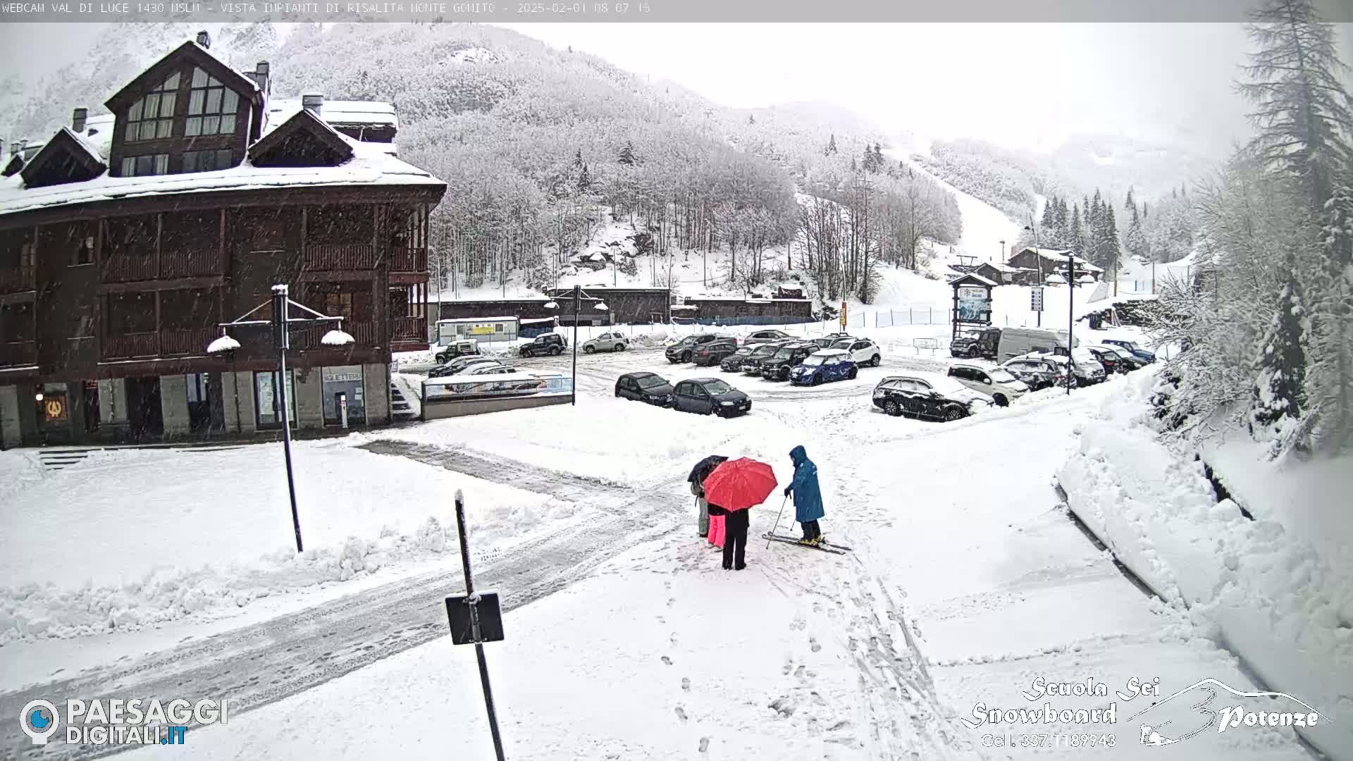 A snow-covered parking lot and a multi-story building are seen in a snowy landscape, with several people standing under umbrellas in the foreground.