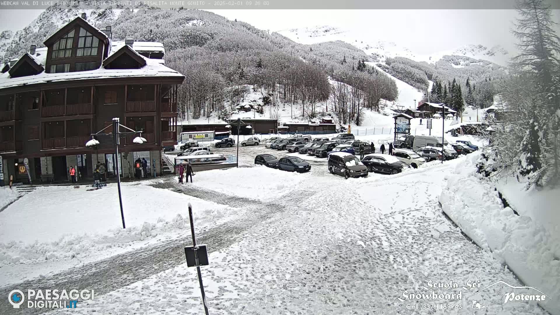 A snow-covered parking lot in front of a multi-story building, with a mountainside covered in snow and trees in the background.