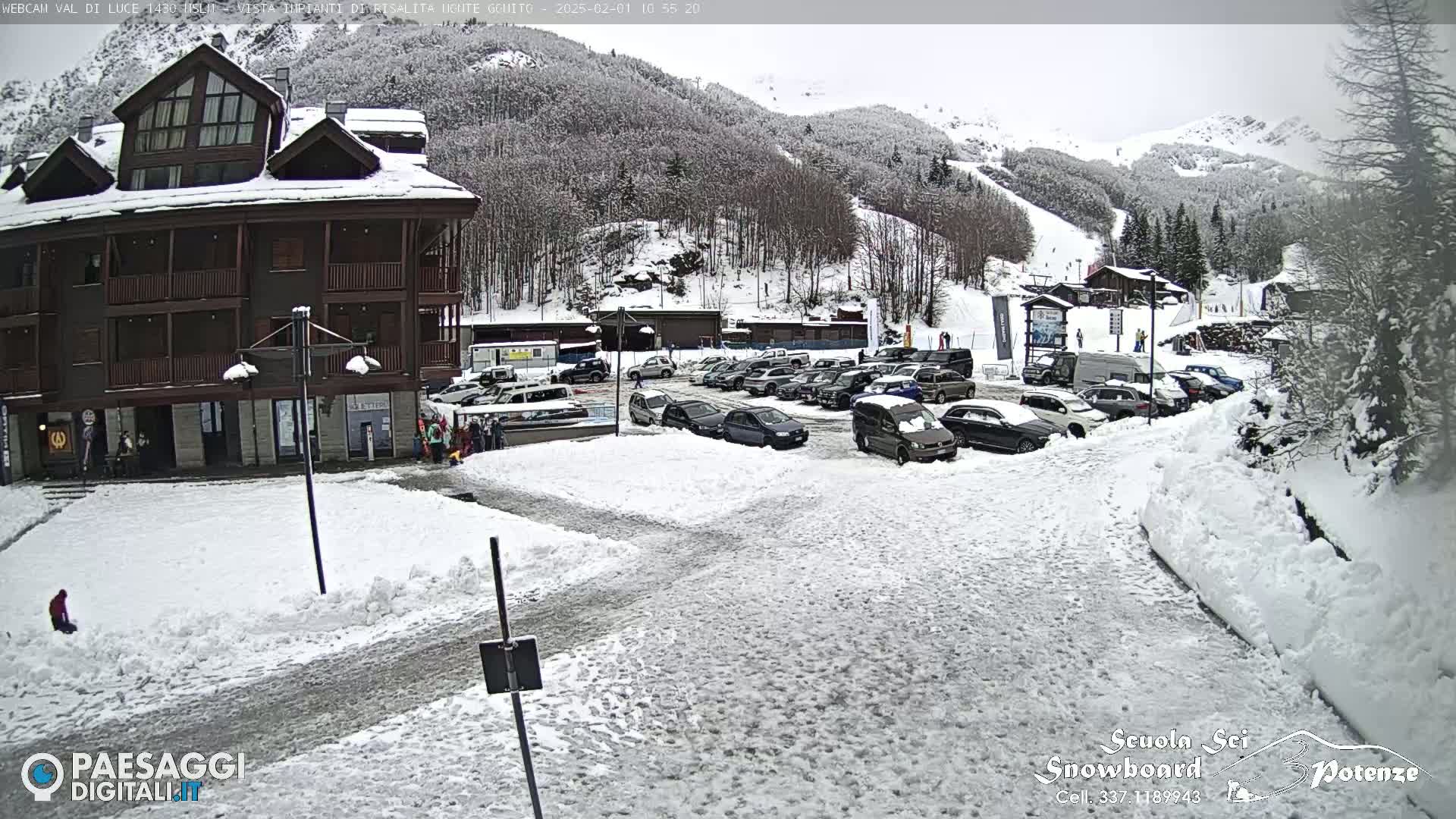 A snow-covered parking lot with cars and a multi-story building is in front of a snow-covered mountainside.