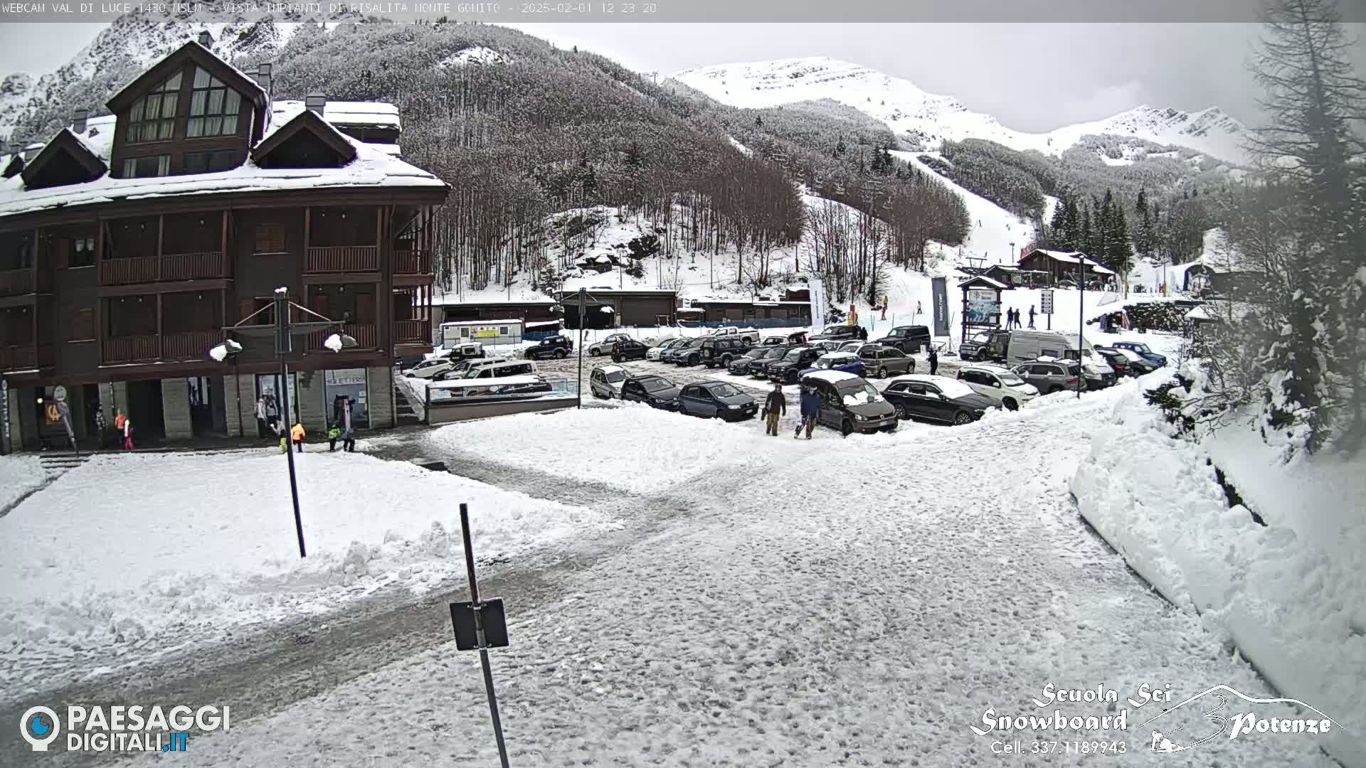 A snow-covered parking lot with cars and a multi-story building is visible, with snow-covered mountains in the background under an overcast sky.