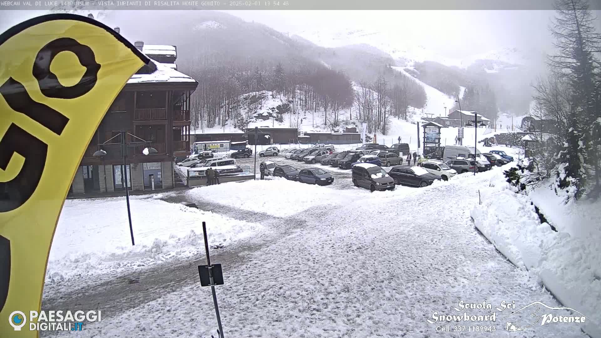 A snow-covered parking lot with cars and a building is visible, with a snowy mountainside and ski slopes in the background under an overcast sky.