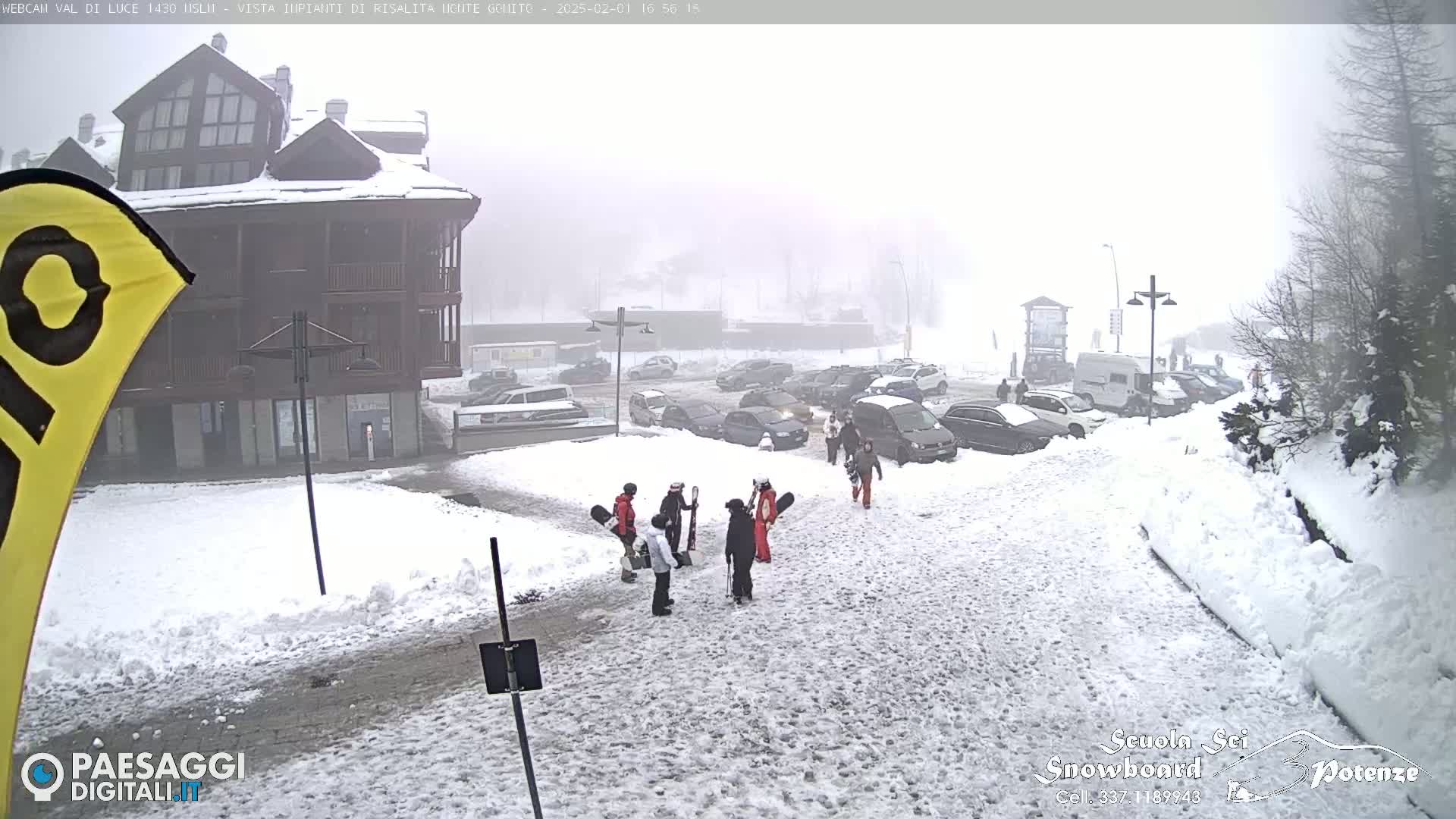 A snow-covered parking lot and street scene, with several people carrying snowboards and skis, are visible in foggy weather conditions.