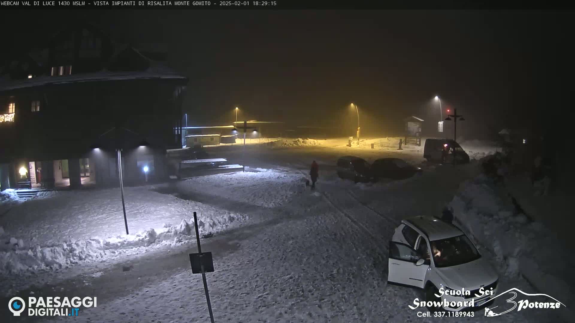 A snow-covered parking lot at night, illuminated by streetlights, with several cars and a building in the background under hazy conditions.