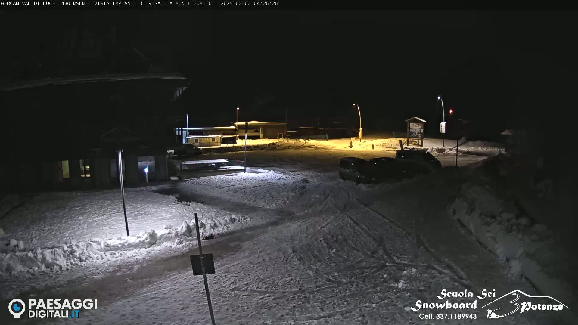 A snow-covered nighttime parking lot with a few cars and buildings under artificial light.