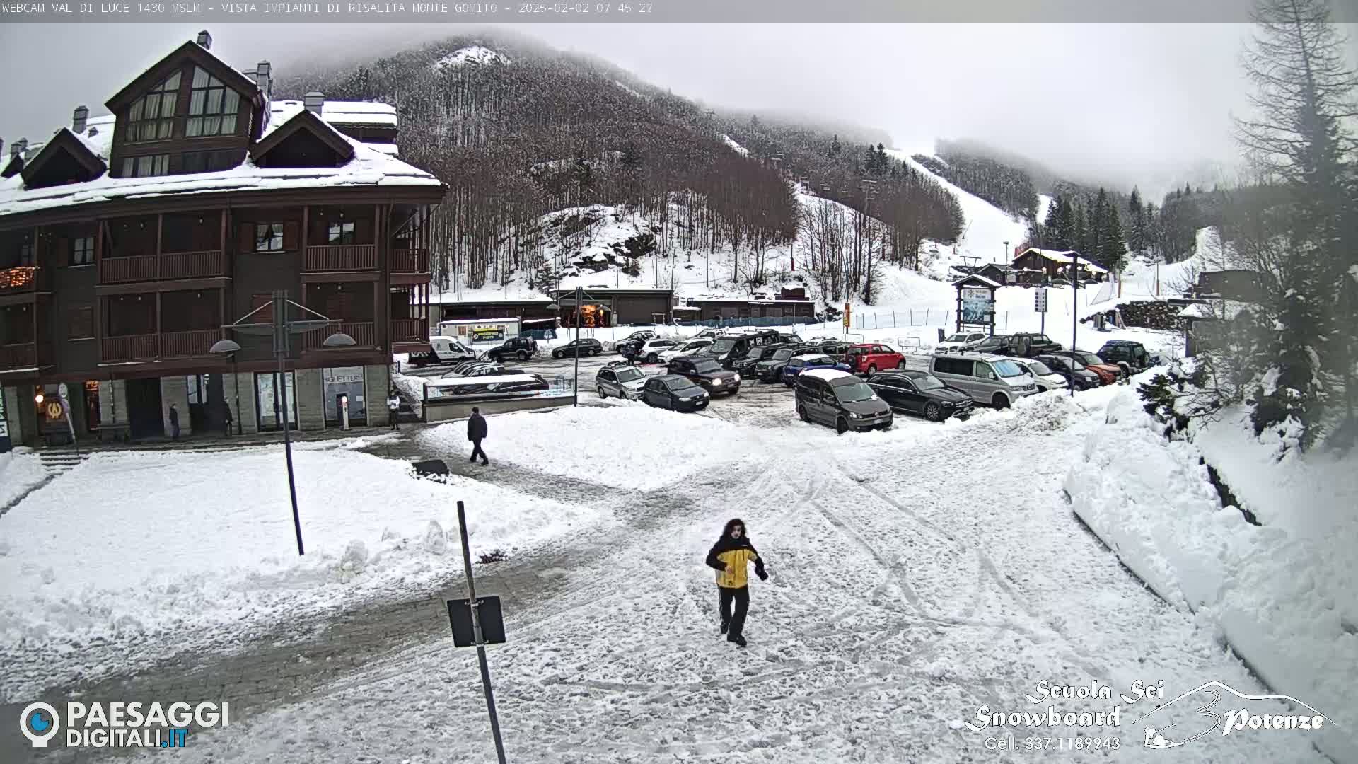 A snow-covered parking lot in front of a multi-story building with a ski slope visible in the background under an overcast sky.
