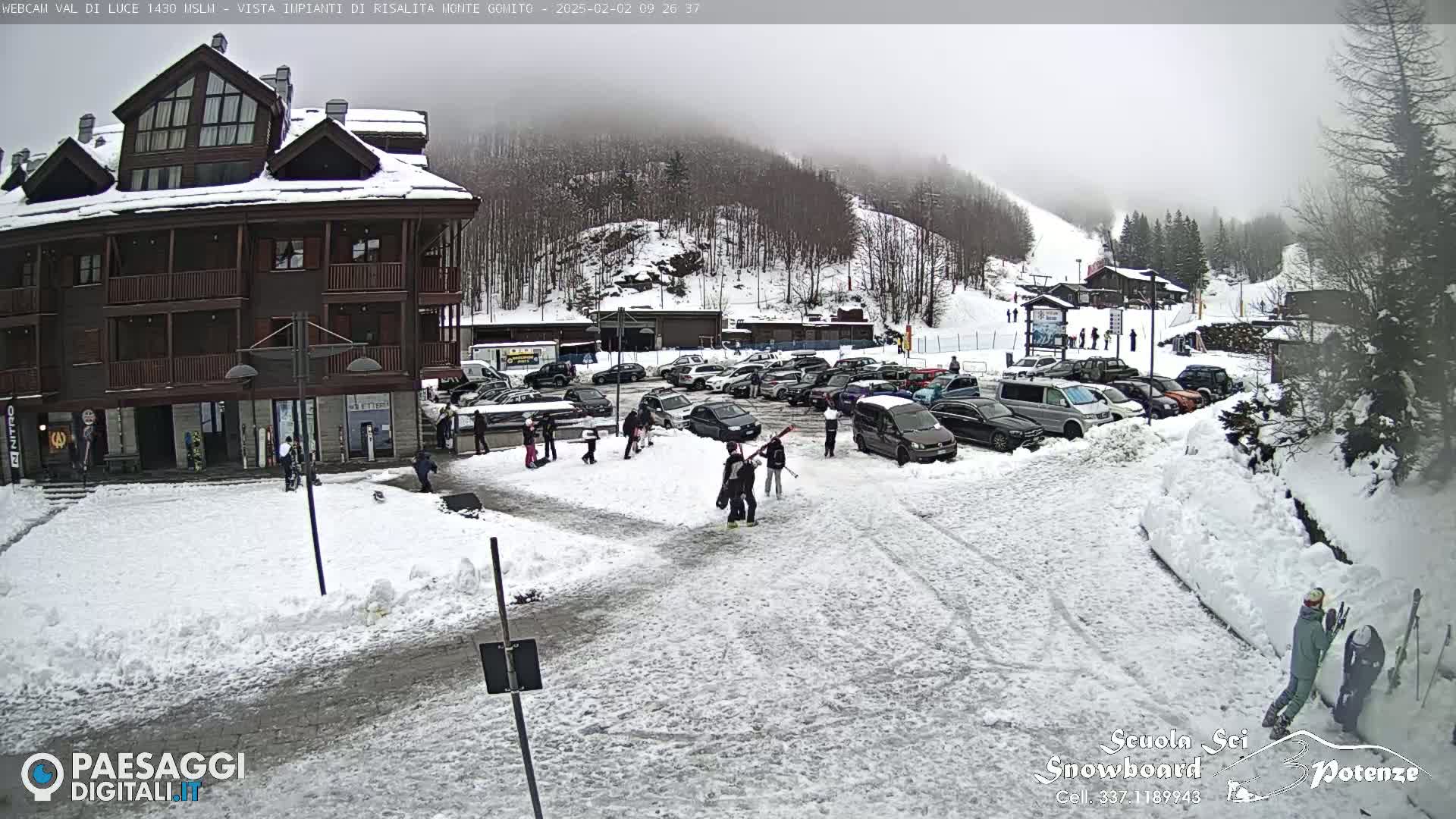 A snow-covered parking lot with cars and several people, situated in front of a multi-story building and a ski slope under a foggy sky.