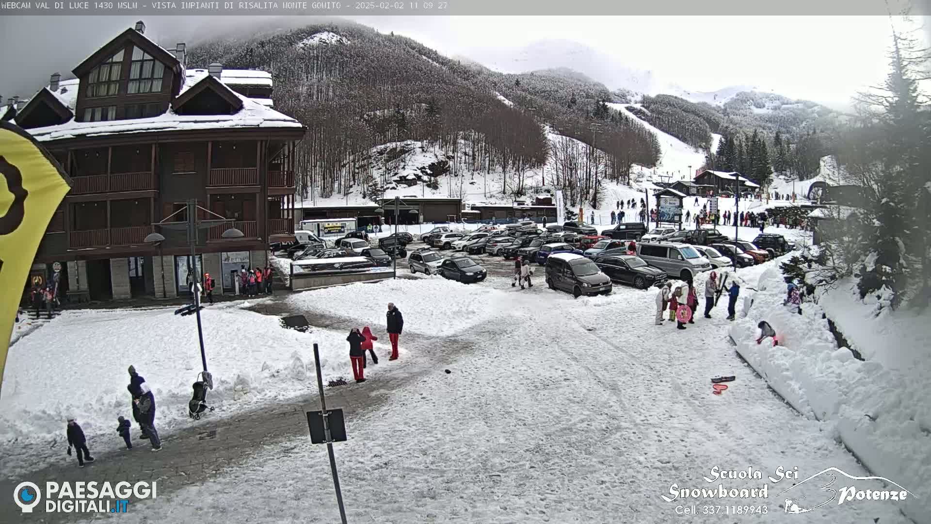A snow-covered parking lot with cars and people near a multi-story building, with a ski slope and snow-covered mountains in the background under an overcast sky.