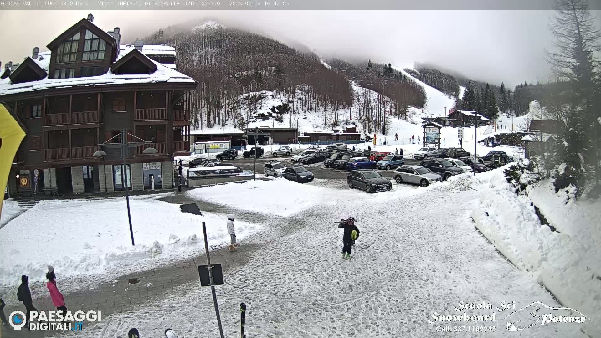 A snow-covered parking lot in front of a multi-story building, with a ski slope visible in the background under a foggy sky.