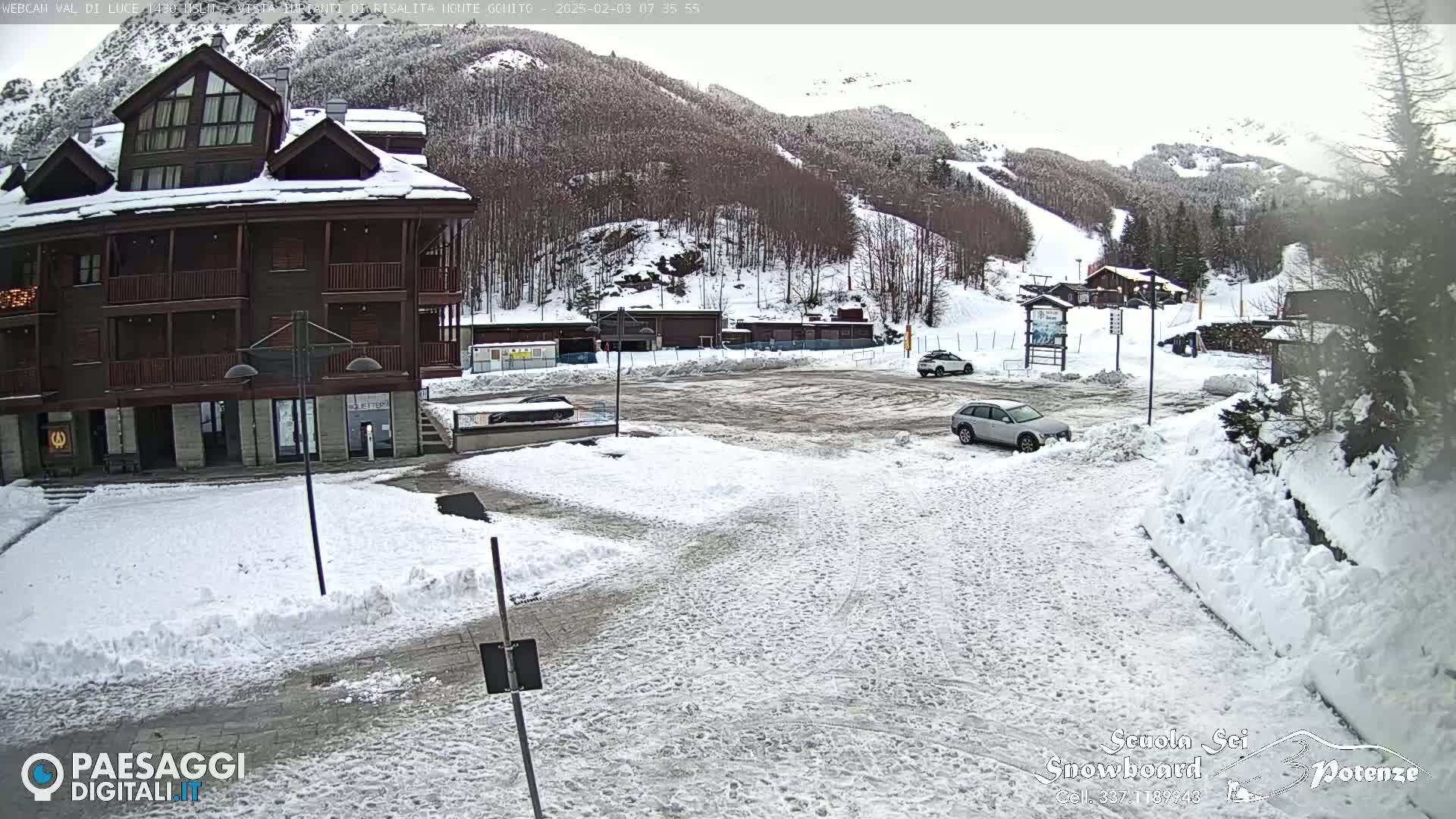 A snow-covered parking lot and roadway are situated in front of a multi-story building, with a mountainside showing ski runs in the background under an overcast sky.