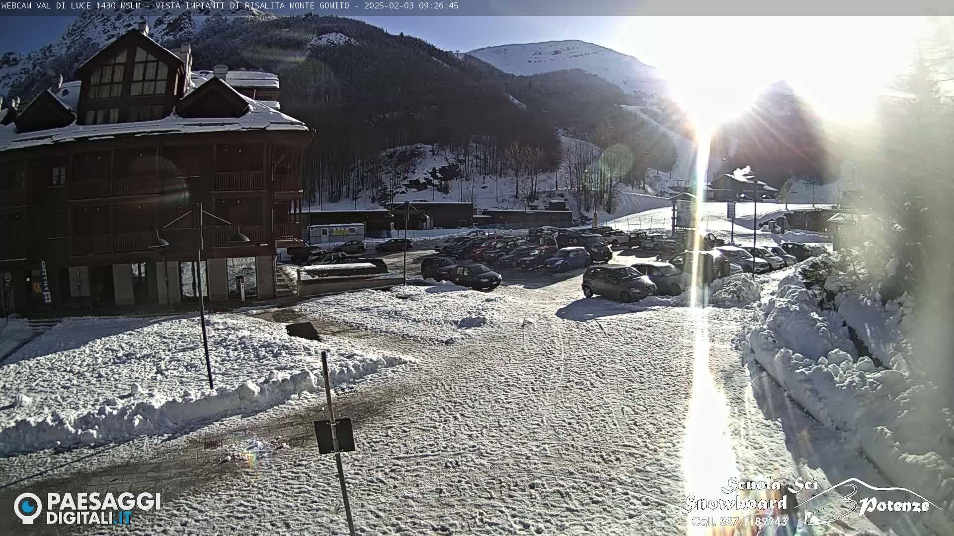 A snow-covered parking lot with cars and a multi-story building sits in front of a sunny, snow-dusted mountainside.