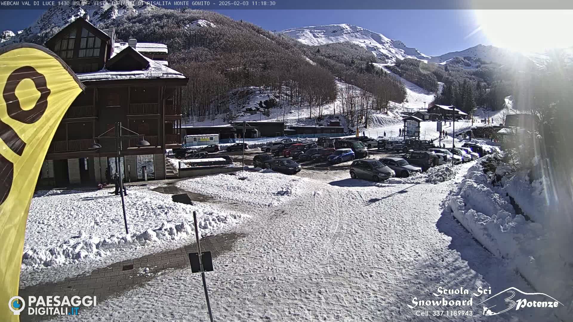A snow-covered parking lot with cars is in front of a multi-story building, with a sunny, snow-dusted mountainside visible in the background.