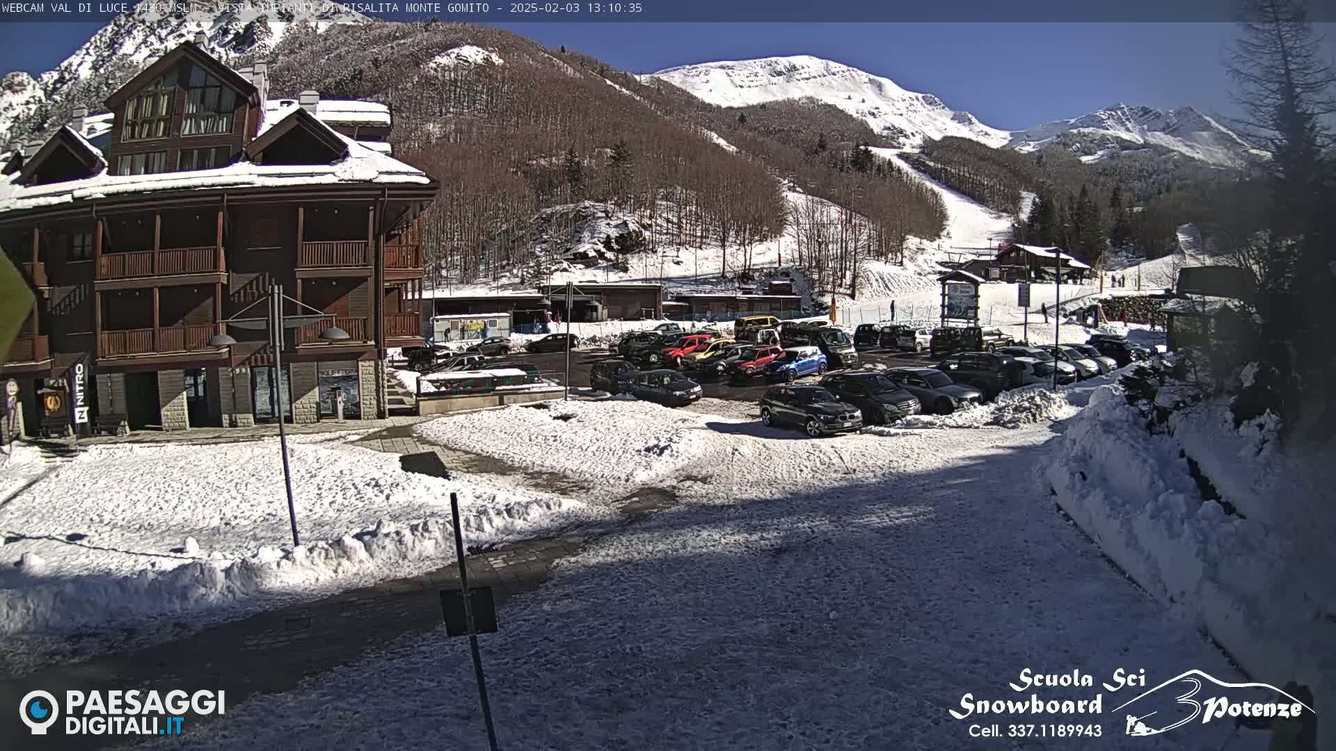 A snow-covered parking lot with cars is situated in front of a multi-story building, all nestled at the base of a sunny, snow-dusted mountain with ski slopes.