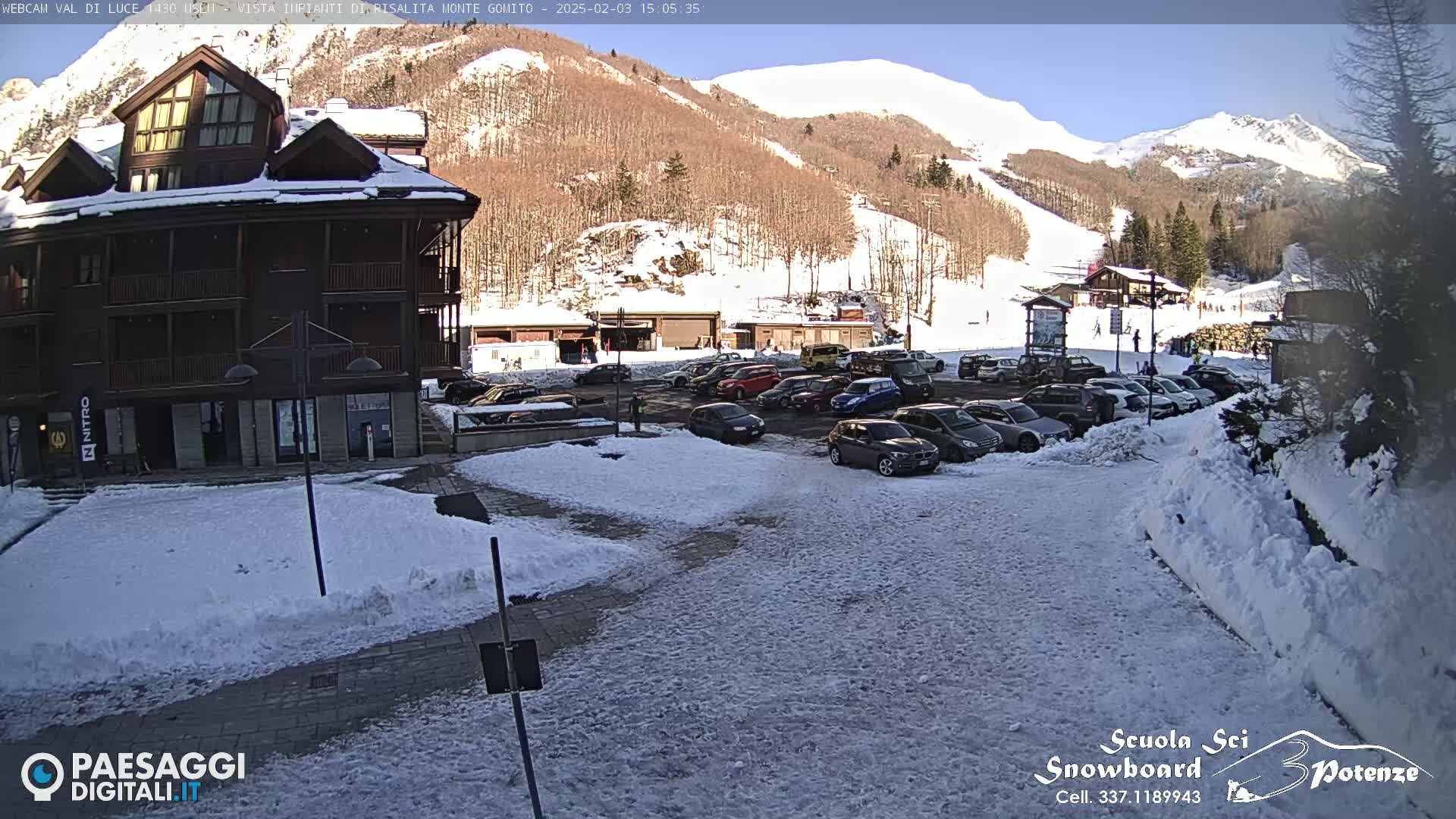 A snow-covered parking lot in front of a multi-story building, with a ski slope and snow-capped mountains in the background under a sunny sky.