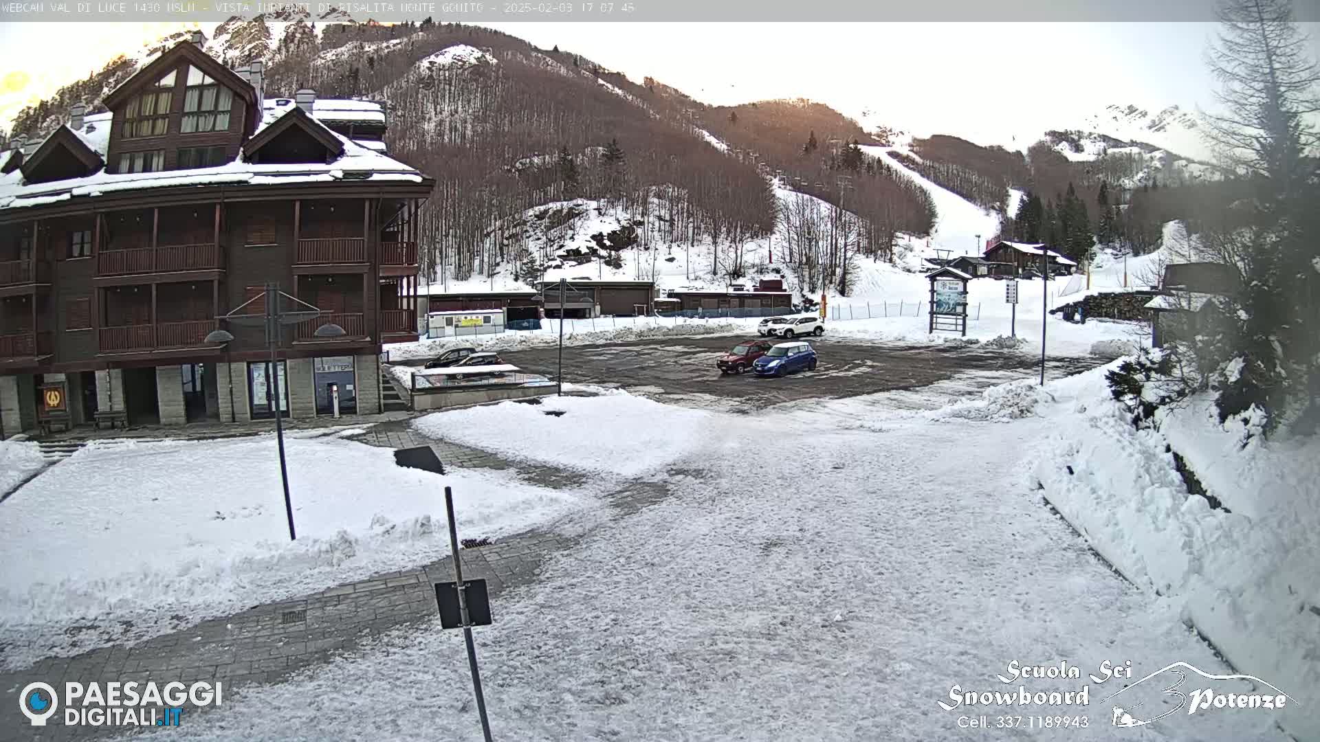 A snow-covered parking lot and plaza are situated in front of a multi-story building, with a mountainside ski slope visible in the background under a partly sunny sky.
