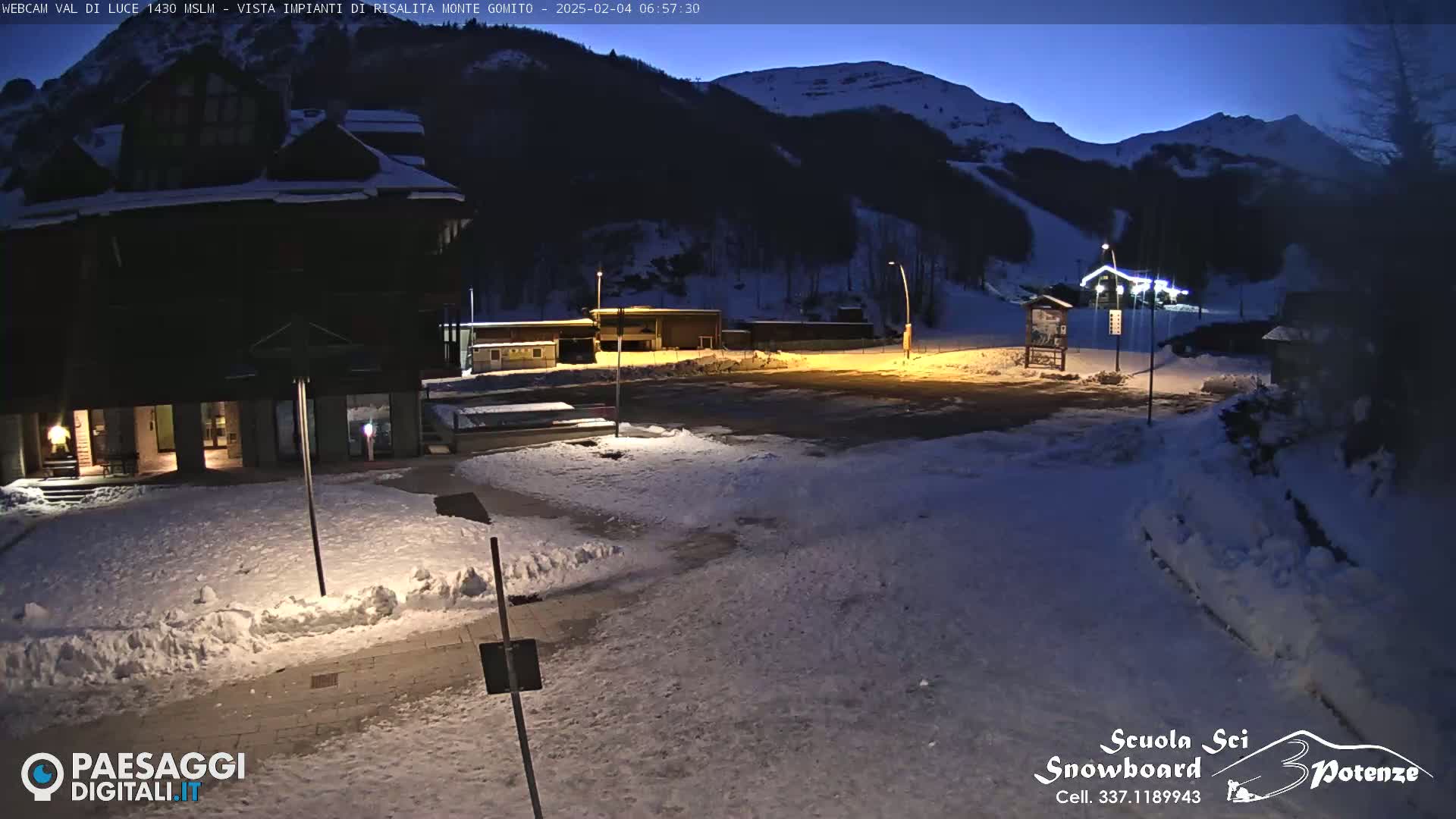 A snow-covered parking lot and buildings are illuminated at night under a twilight sky, with mountains in the background.
