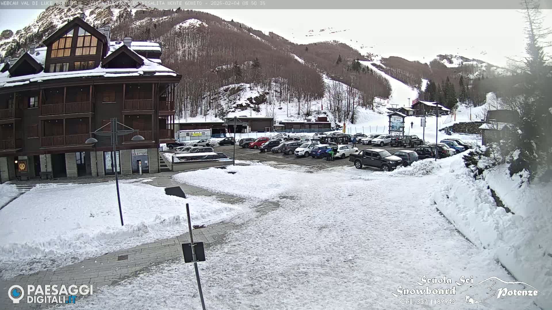 A snow-covered parking lot with cars in front of a multi-story brown building, nestled against a snow-dusted mountainside.