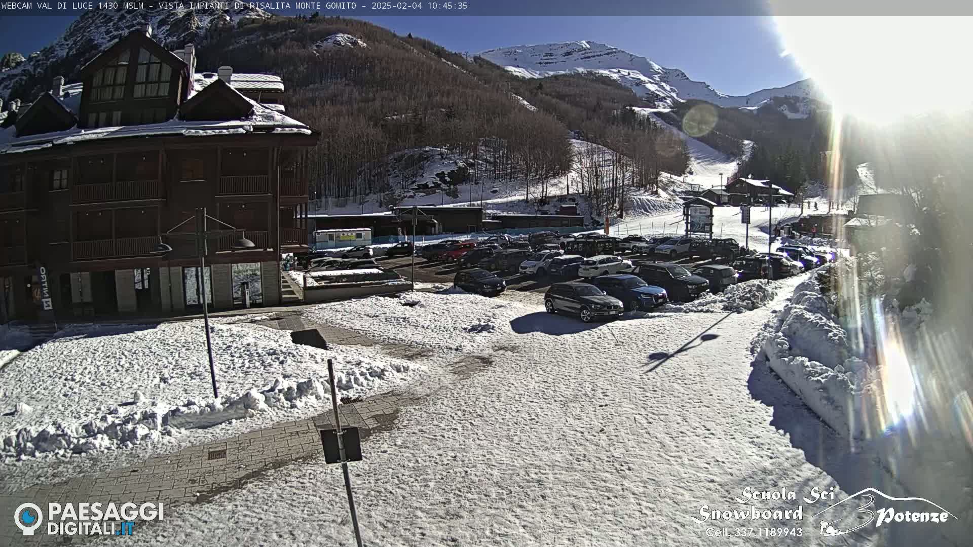 A snow-covered parking lot with cars and a multi-story building is seen on a sunny day with mountains in the background.