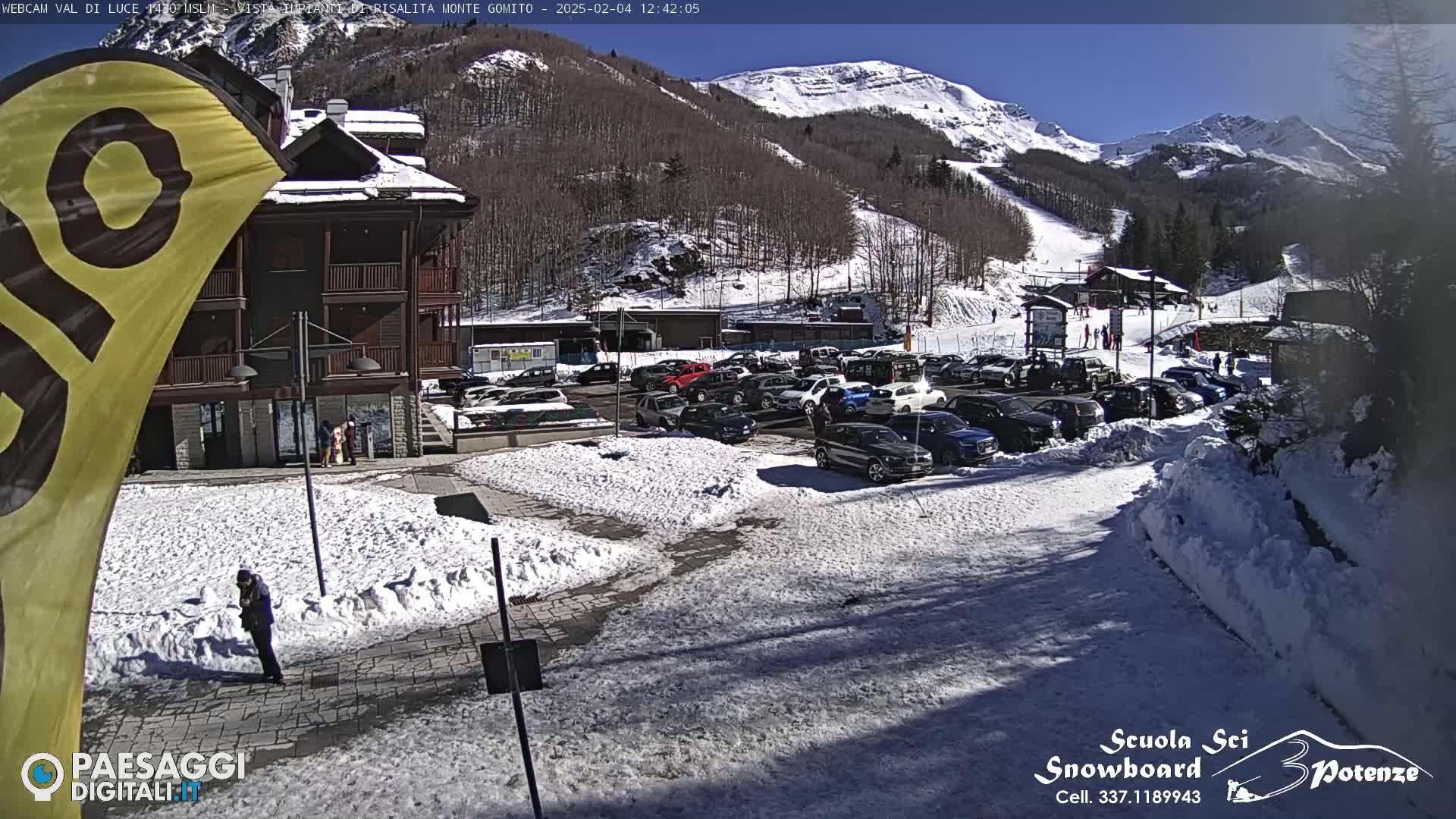 A sunny day in a snow-covered mountain resort parking lot, with a building and ski slope in the background.