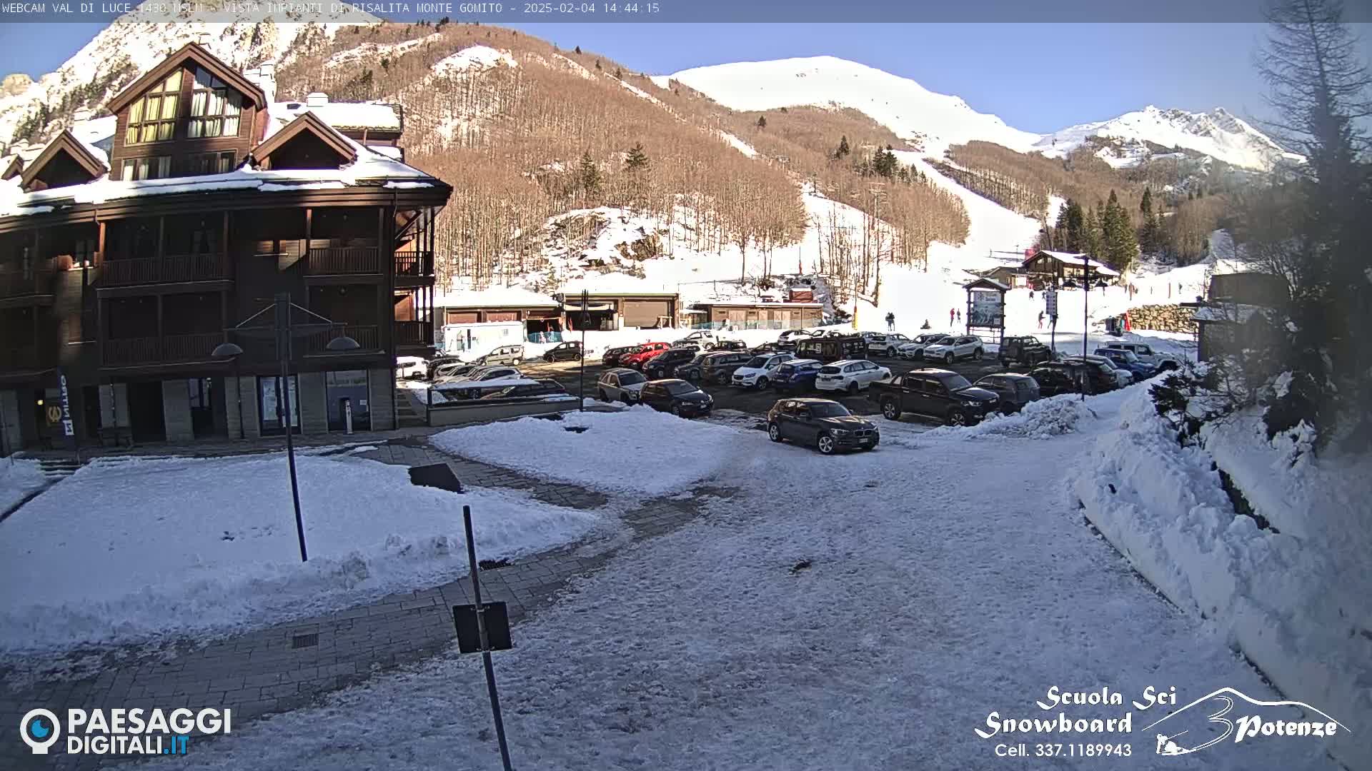 A snow-covered parking lot is seen in front of a multi-story building, with a sunny, snow-dusted mountainside in the background.