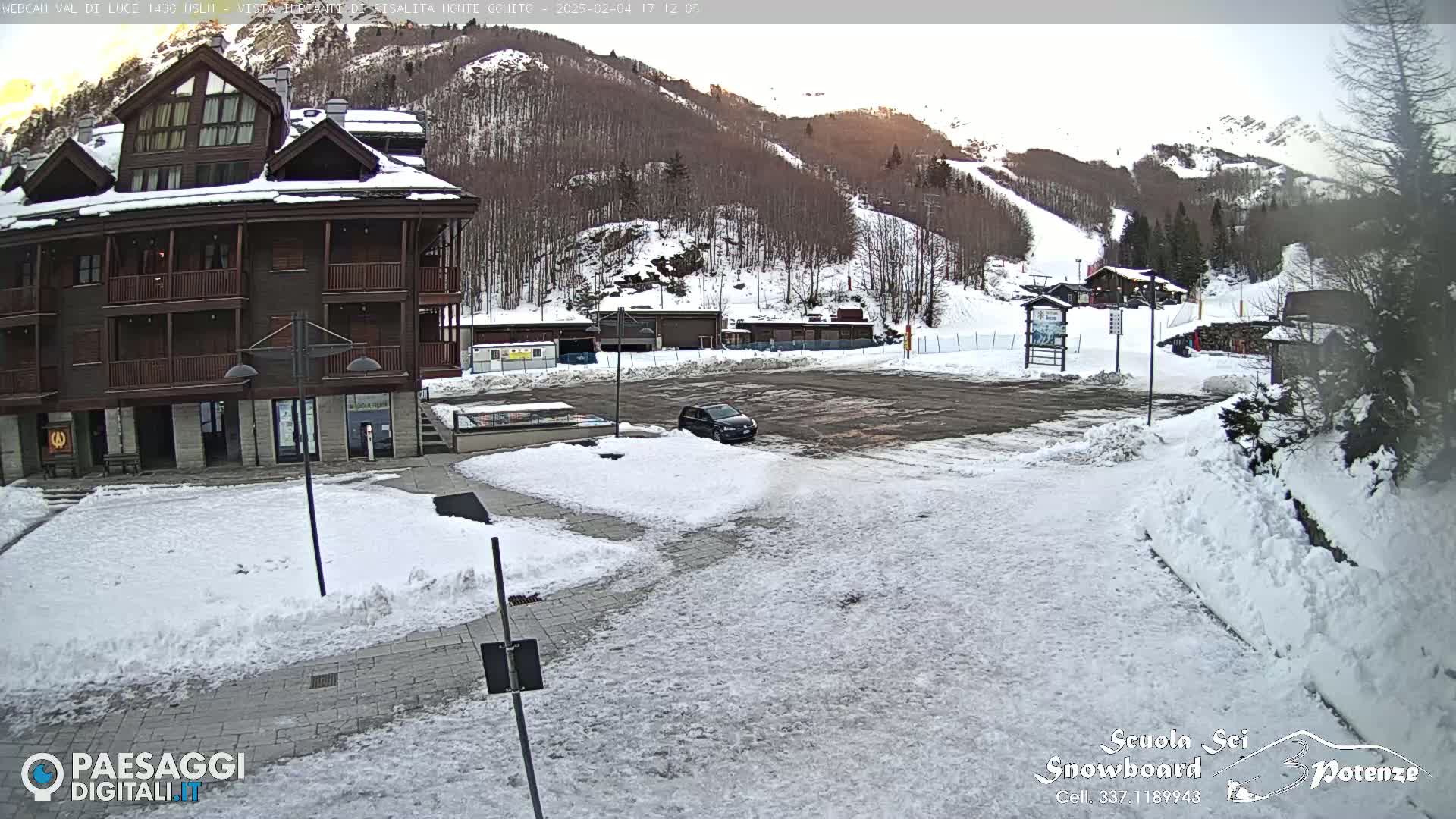 A snow-covered parking lot sits in front of a multi-story brown building, with a mountain ski slope visible in the background under a partly sunny sky.