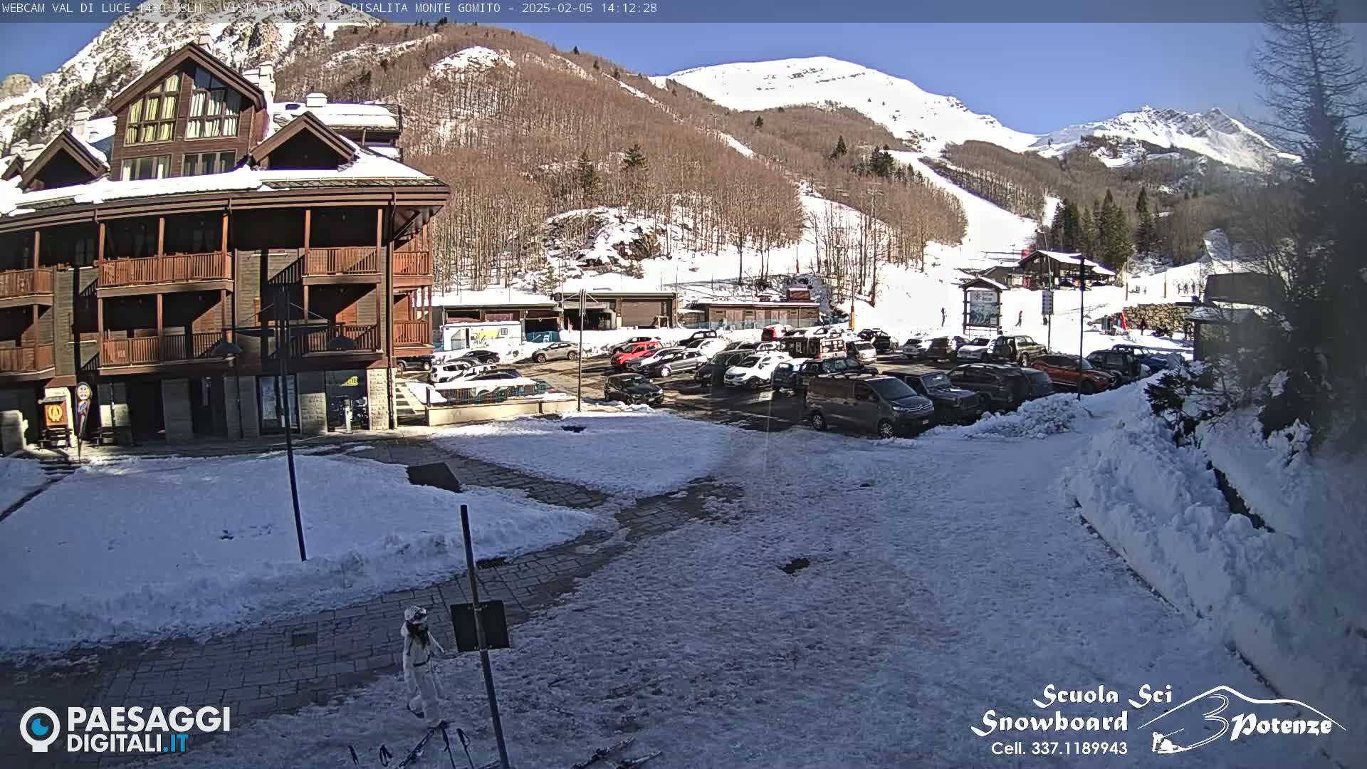 A snow-covered parking lot is in front of a multi-story building, with a sunny, snow-dusted mountain in the background.