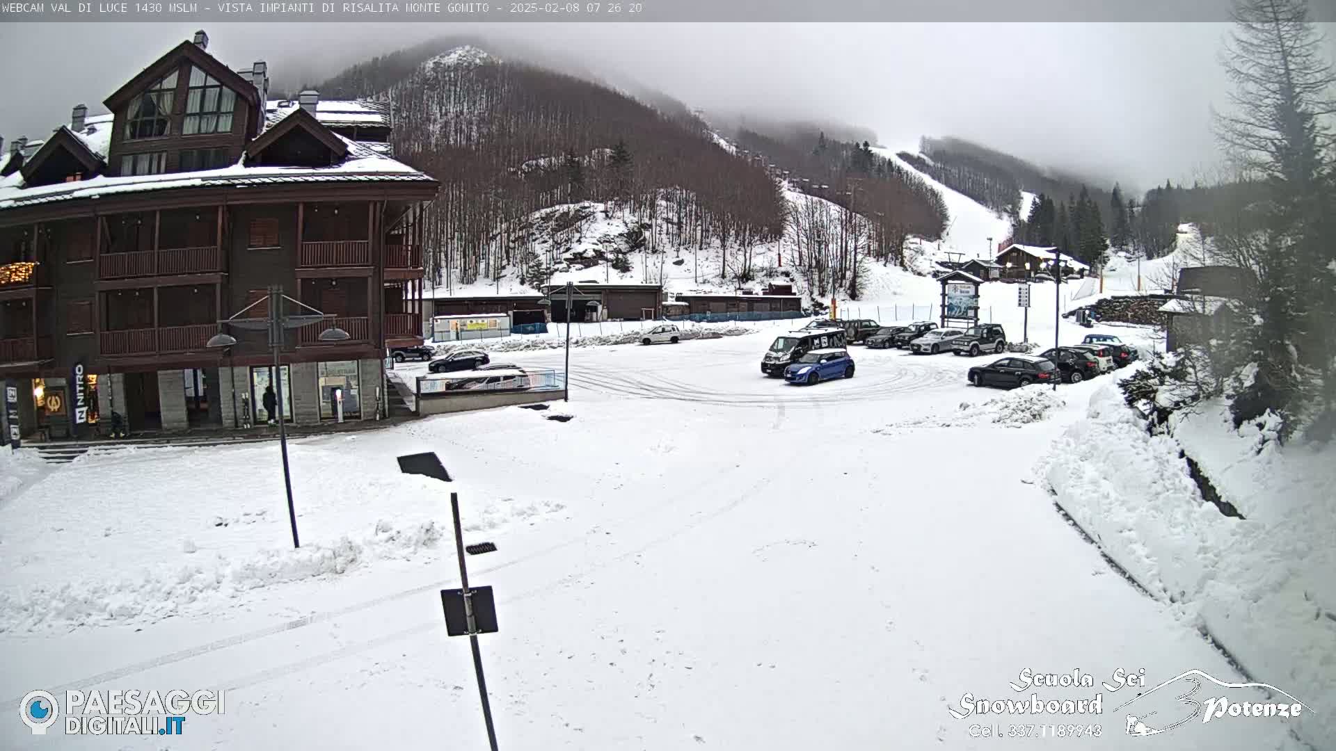 A snow-covered parking lot with several cars sits in front of a multi-story building, with a snow-covered mountain in the background under overcast skies.
