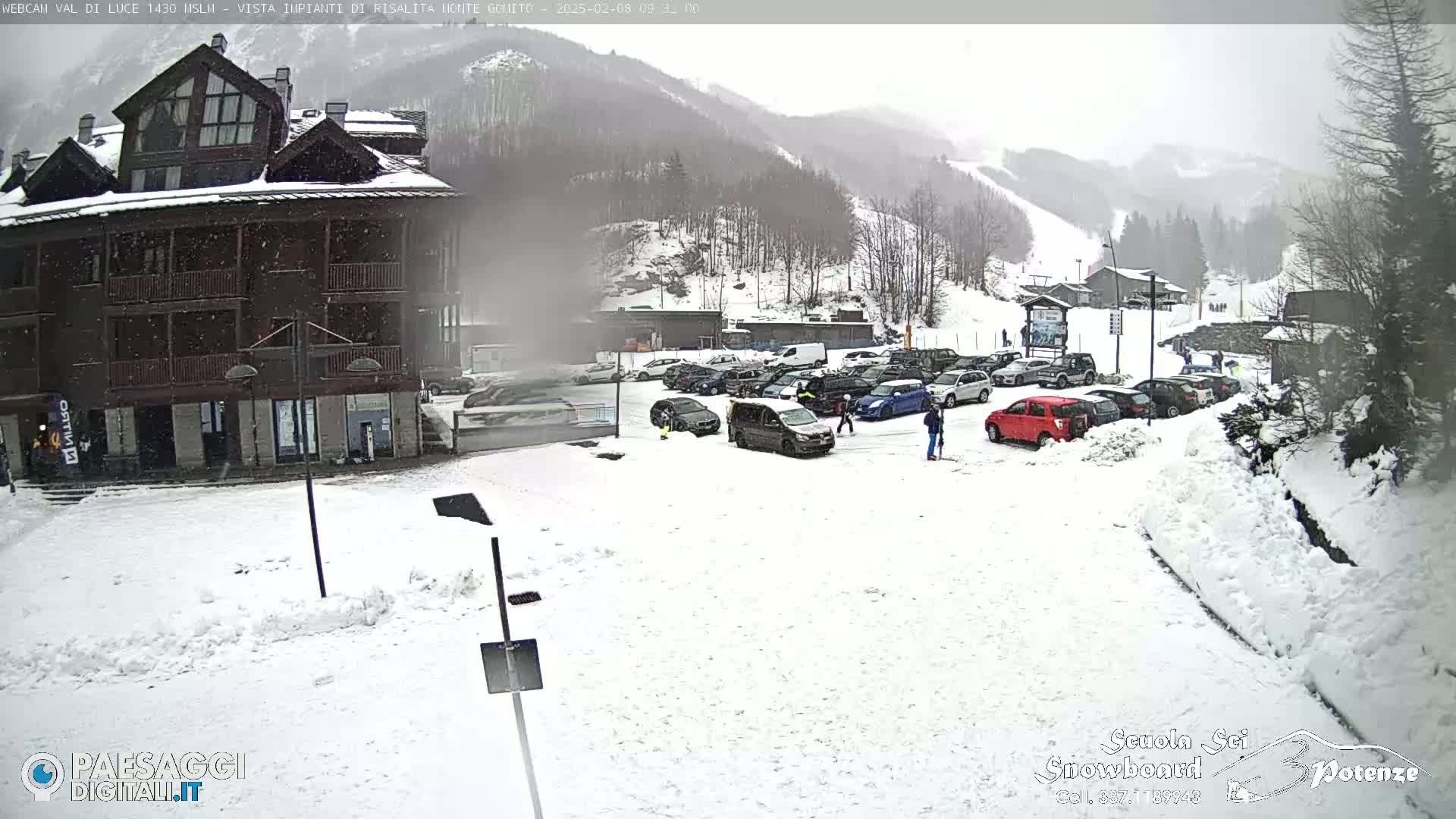 A snow-covered parking lot with cars and a multi-story building is visible in the foreground, with a snow-dusted mountainside in the background under overcast skies.