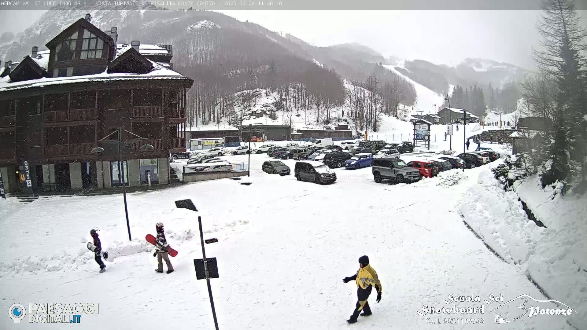 A snow-covered parking lot with cars and a few people, including two snowboarders, sits in front of a multi-story building with snowy mountains and ski slopes in the background during a light snowfall.
