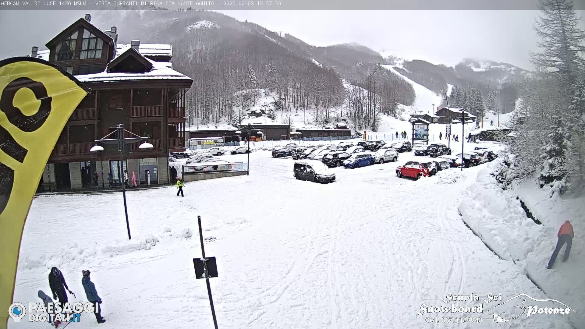 A snow-covered parking lot with several cars and a multi-story building is seen under an overcast sky, with a ski slope visible in the background.