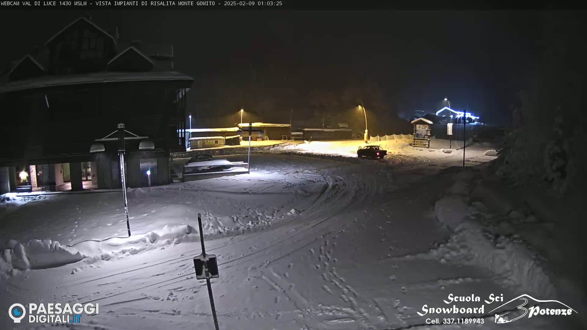 A snow-covered parking lot and roadway at night, illuminated by streetlights, with a dark-colored vehicle visible in the distance.