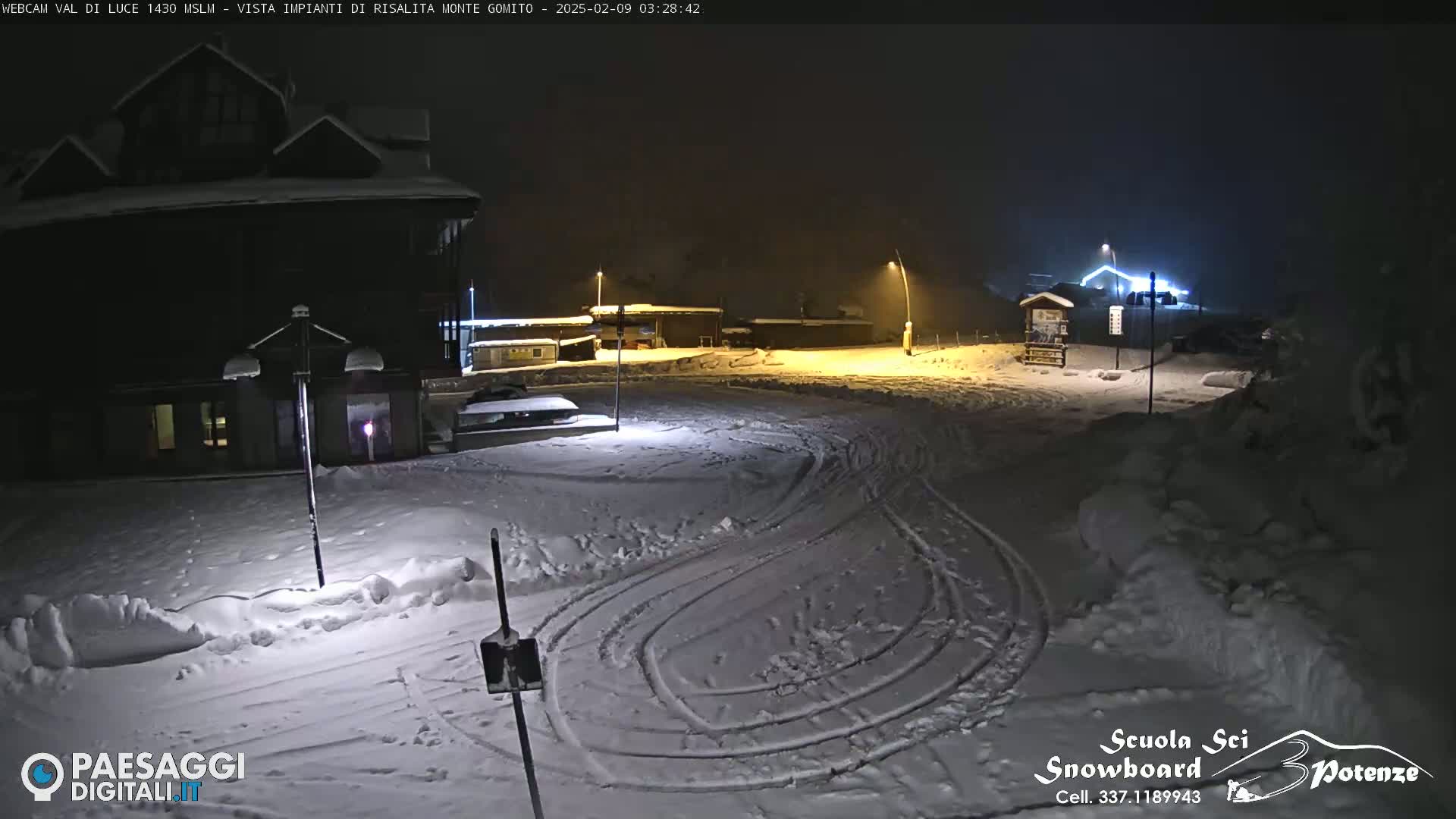 A snow-covered parking lot at night is illuminated by several streetlights, with tire tracks visible in the snow and a building in the background.