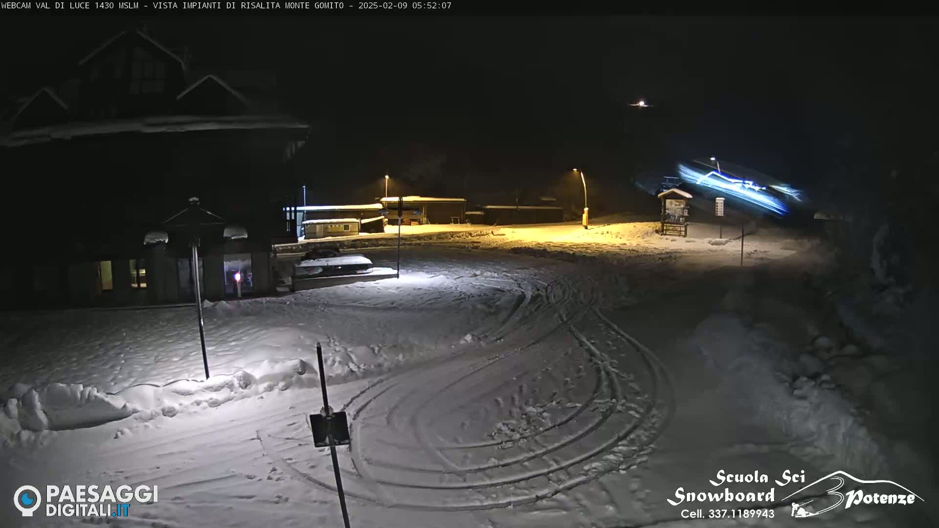 A snow-covered parking lot at night, illuminated by streetlights, shows tire tracks curving through the snow, with a building and a blurry light trail in the background.