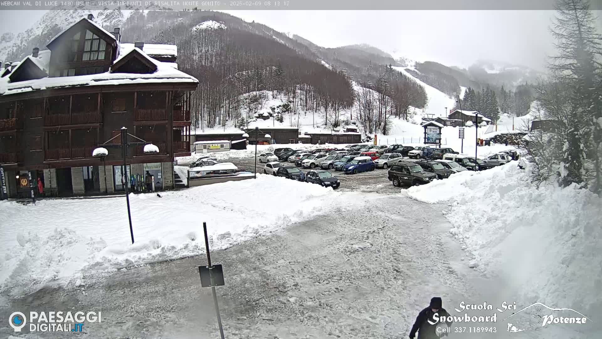 A snow-covered parking lot with numerous cars is situated in front of a multi-story building, with a snow-covered mountainside and ski slopes in the background under an overcast sky.