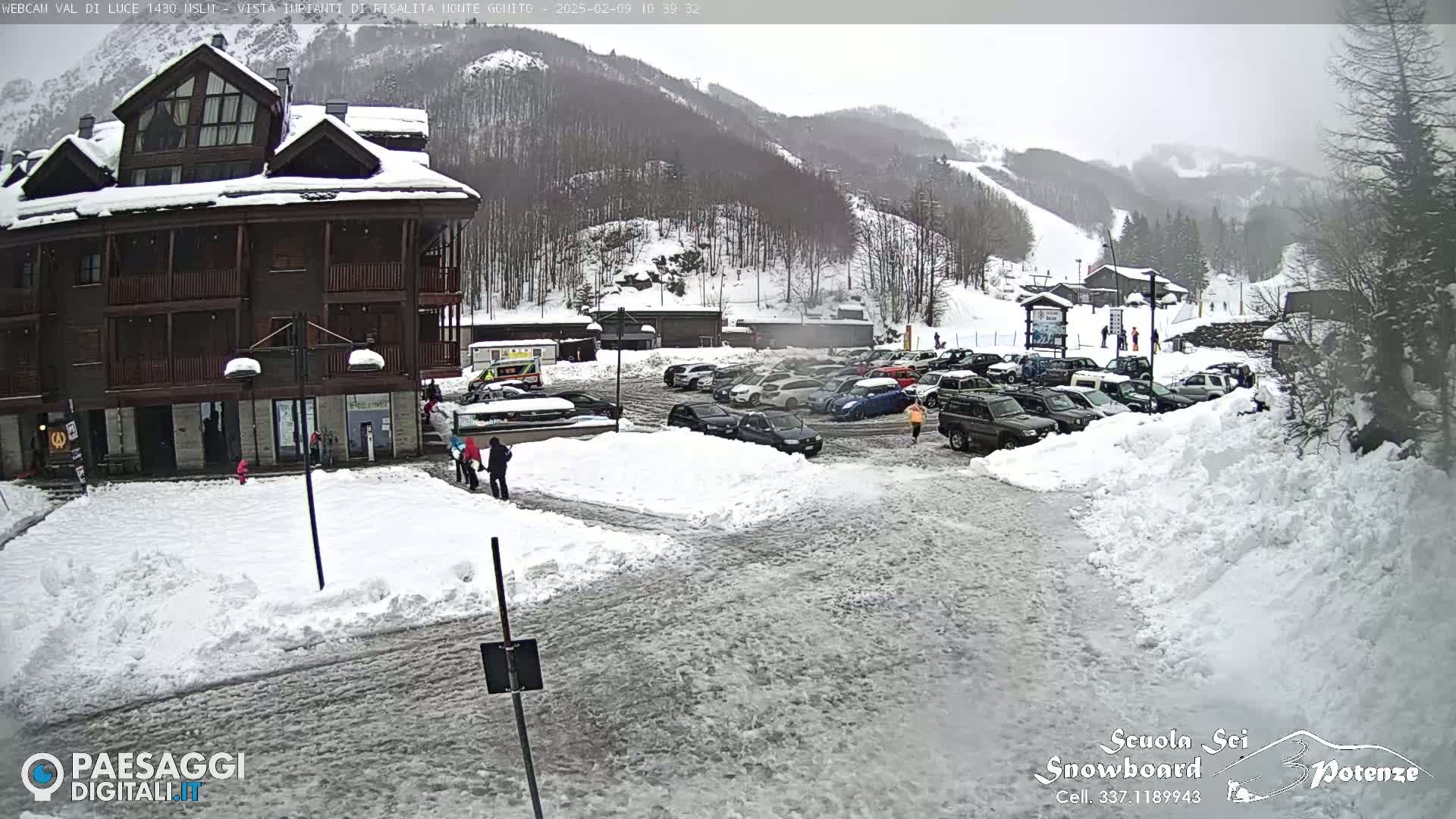 A snow-covered parking lot with cars and a multi-story building sits at the base of a snow-dusted mountain on an overcast day.