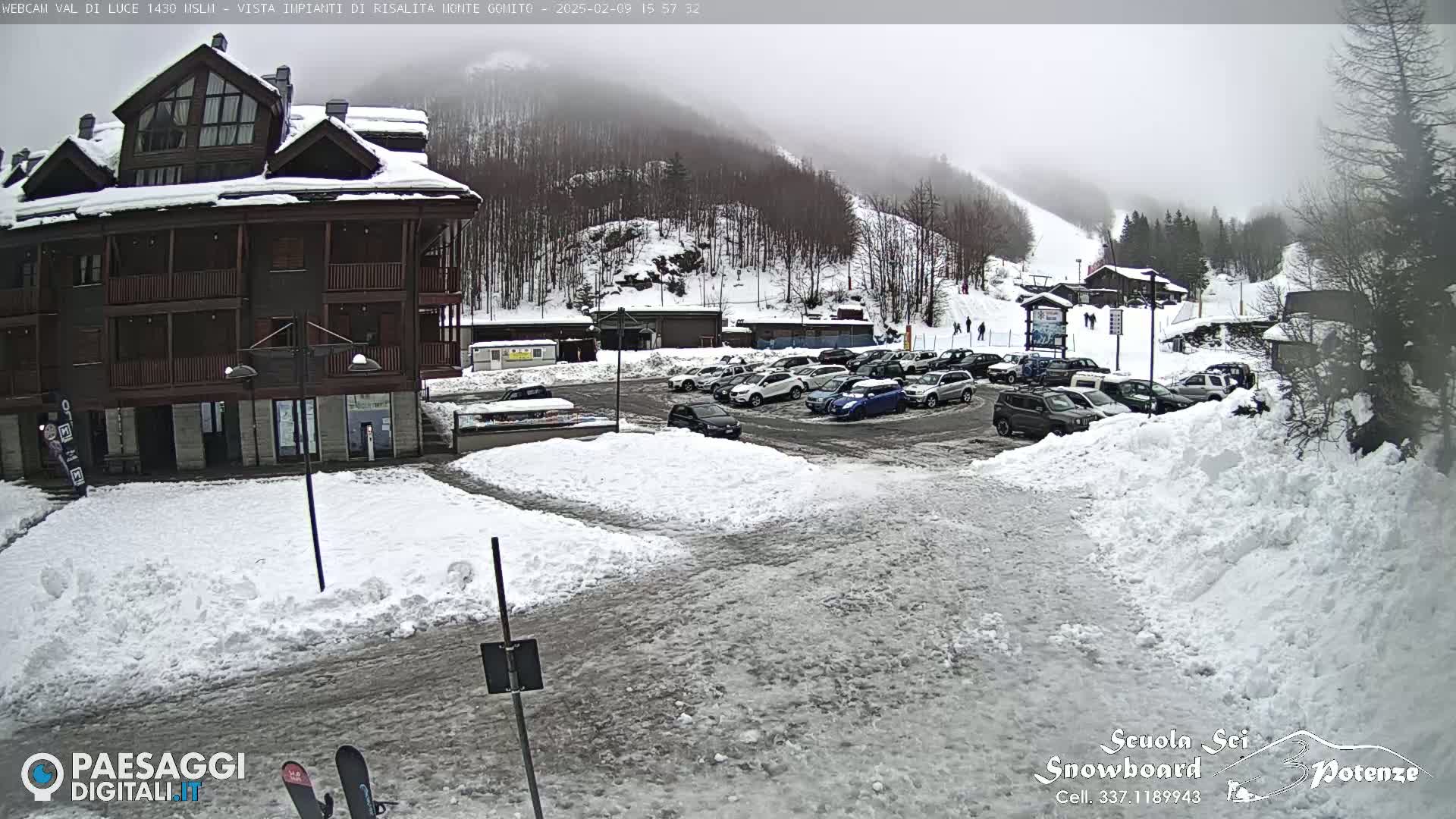 A snow-covered parking lot with cars and a multi-story building is visible, with a foggy, snowy mountainside in the background.