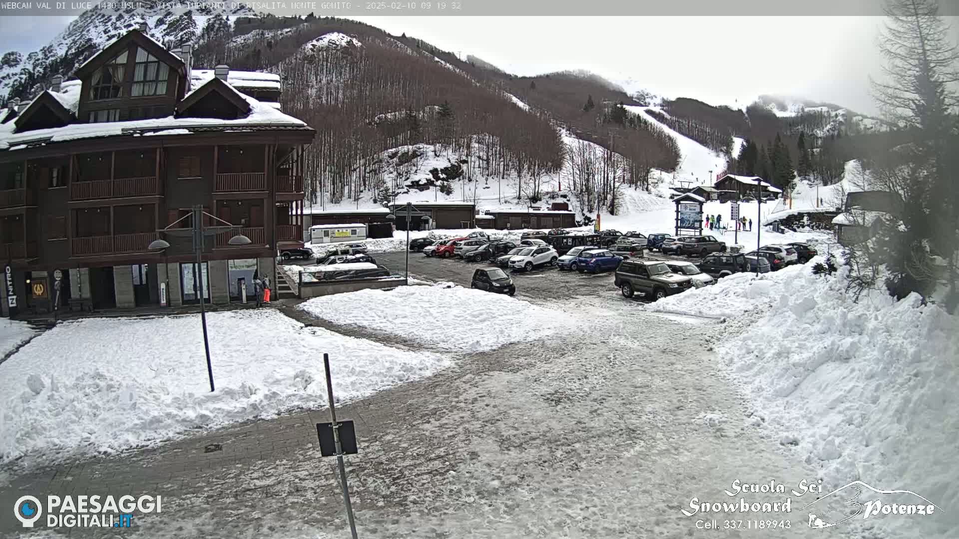 A snow-covered parking lot with cars sits in front of a multi-story building, with a snowy mountain slope visible in the background under an overcast sky.