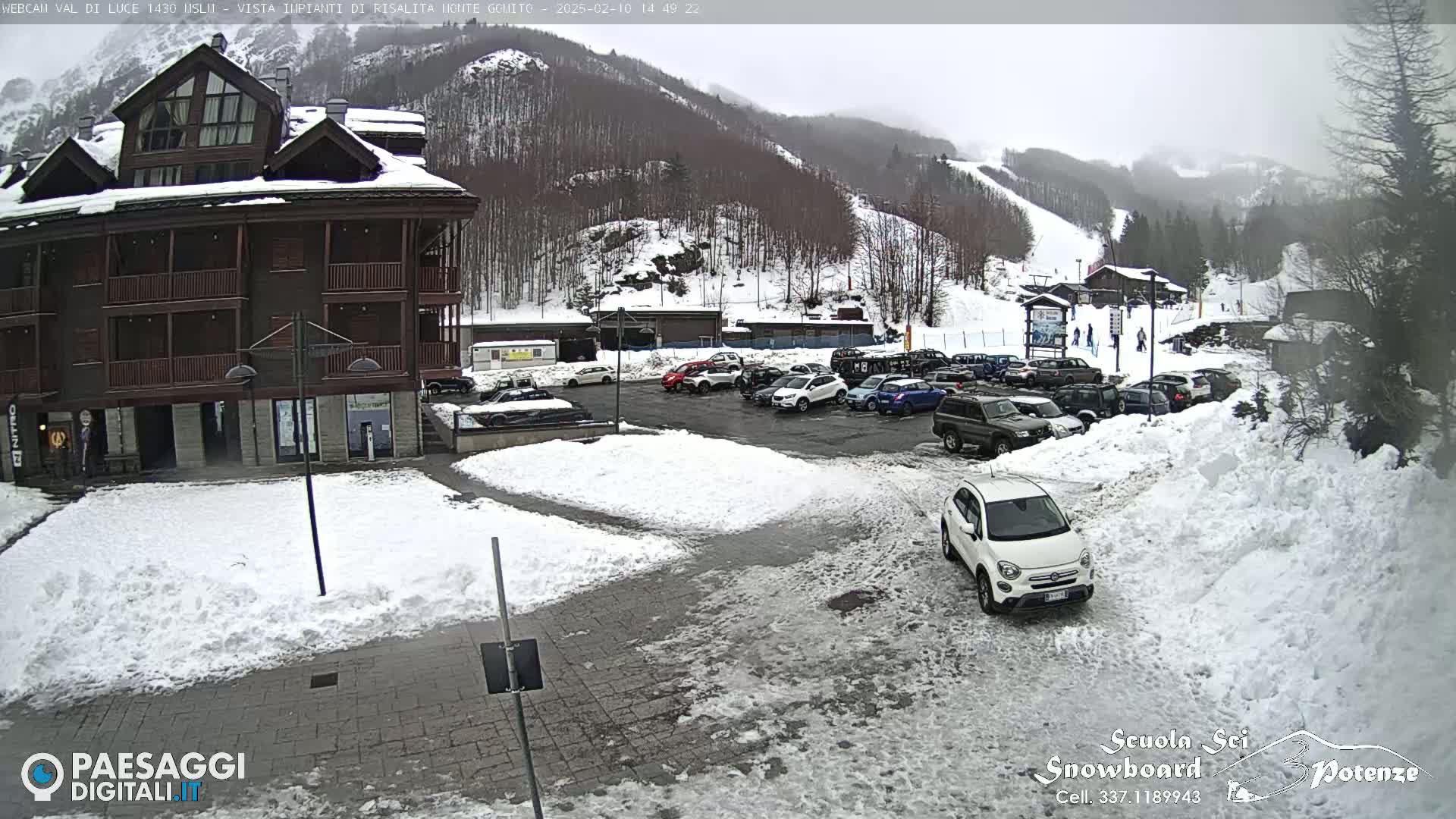 A snow-covered parking lot with cars and a multi-story building is situated in front of a snow-covered mountain slope on an overcast day.