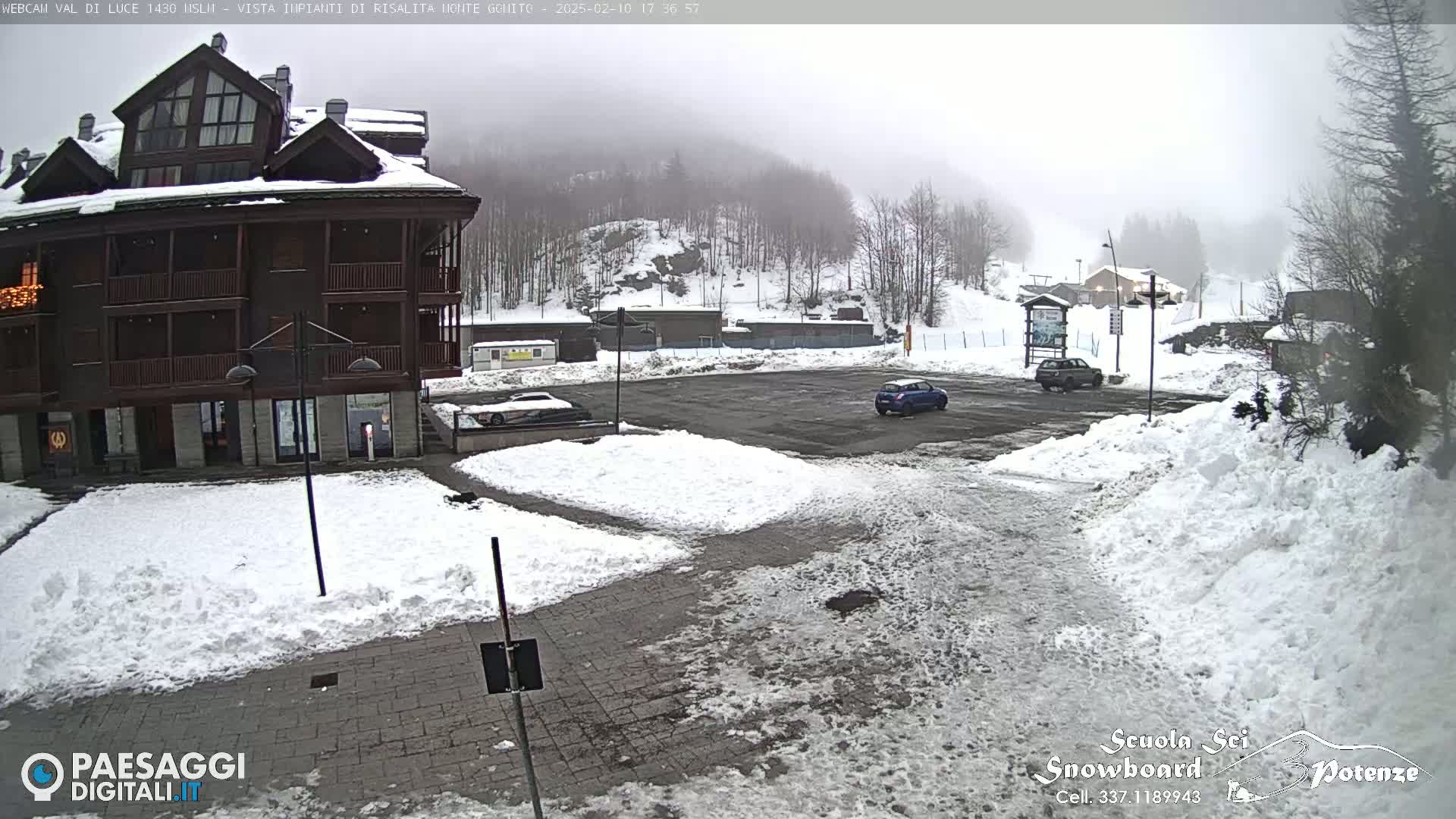 A snow-covered parking lot sits in front of a multi-story brown building, with a foggy, snowy mountainside in the background.