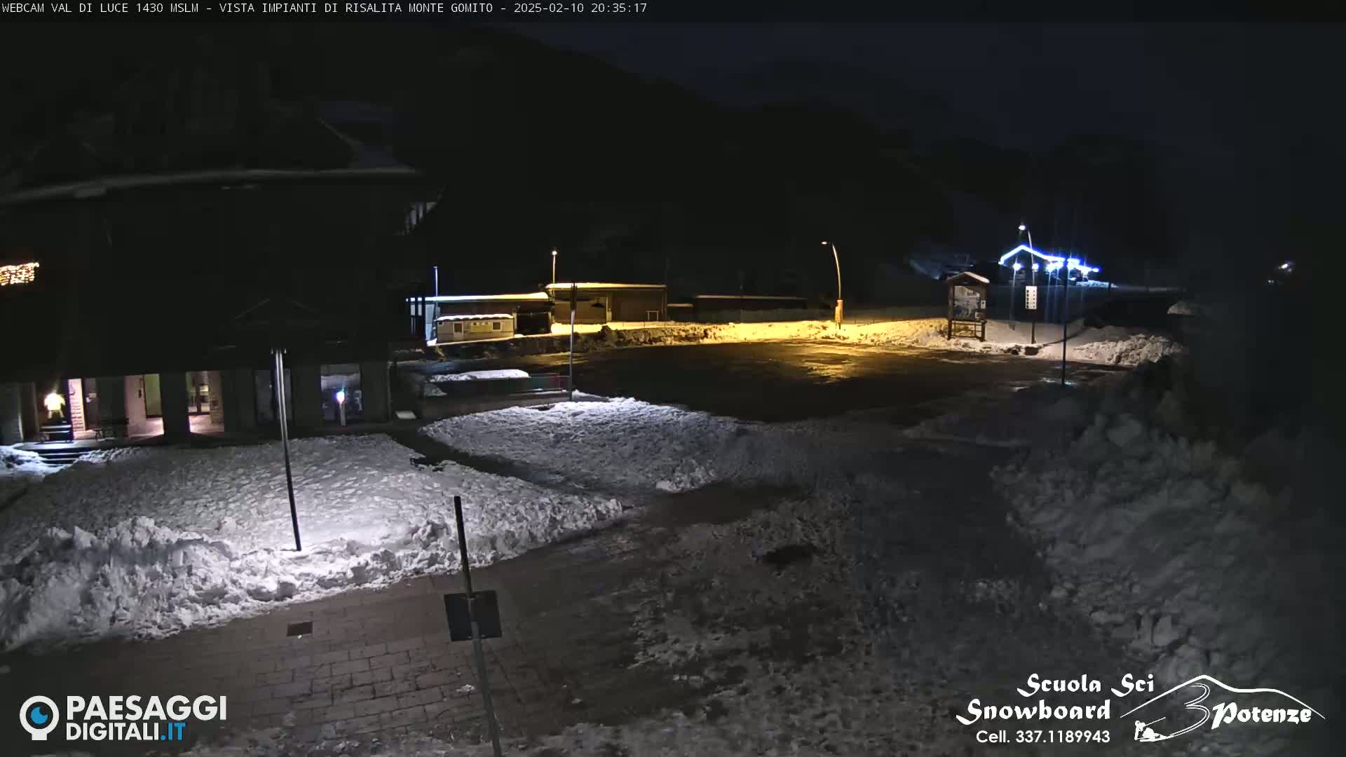 A snow-covered nighttime scene shows a building and a parking lot illuminated by lights, with a mountain in the background.