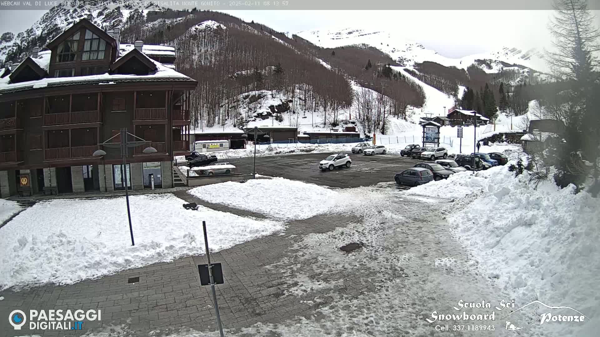 A snow-covered parking lot with several cars is in front of a brown multi-story building, with a snowy mountainside in the background under an overcast sky.