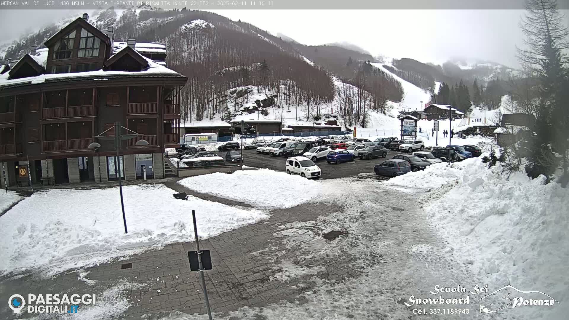A snow-covered parking lot with cars and a multi-story building is visible, with a snowy mountainside and ski slope in the background under an overcast sky.