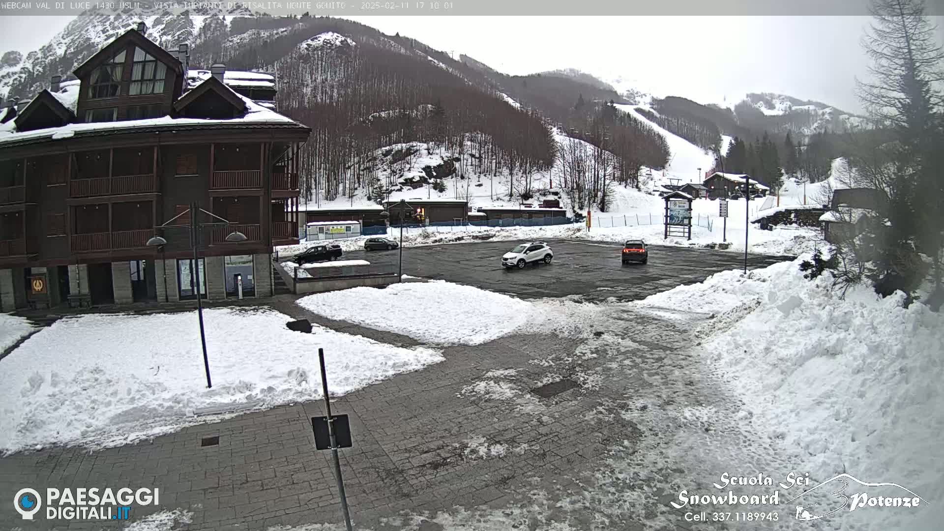 A snow-covered parking lot with a few cars sits in front of a multi-story brown building, with snow-covered mountains and ski slopes in the background under an overcast sky.