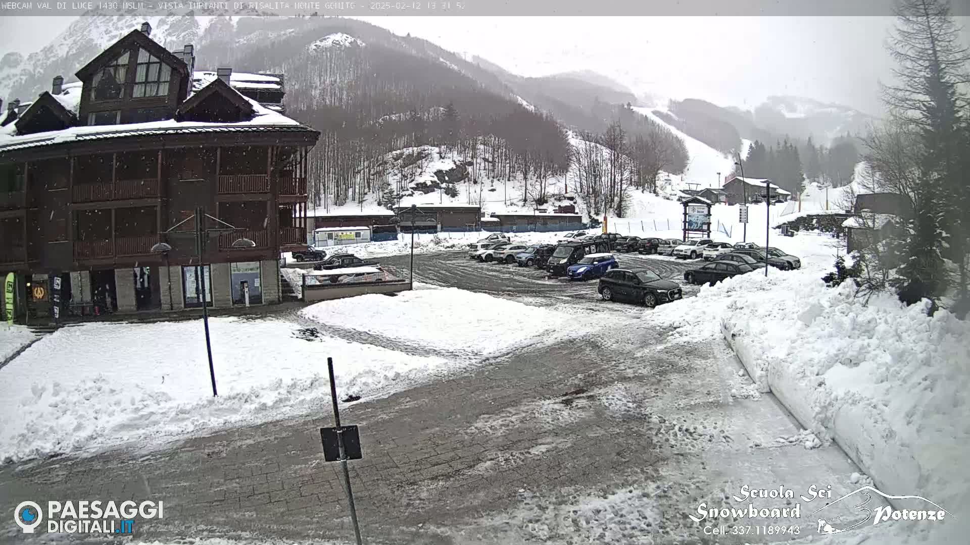 A snow-covered parking lot with several cars is situated in front of a multi-story wooden building, with snow-covered mountains and ski slopes visible in the background under an overcast sky.