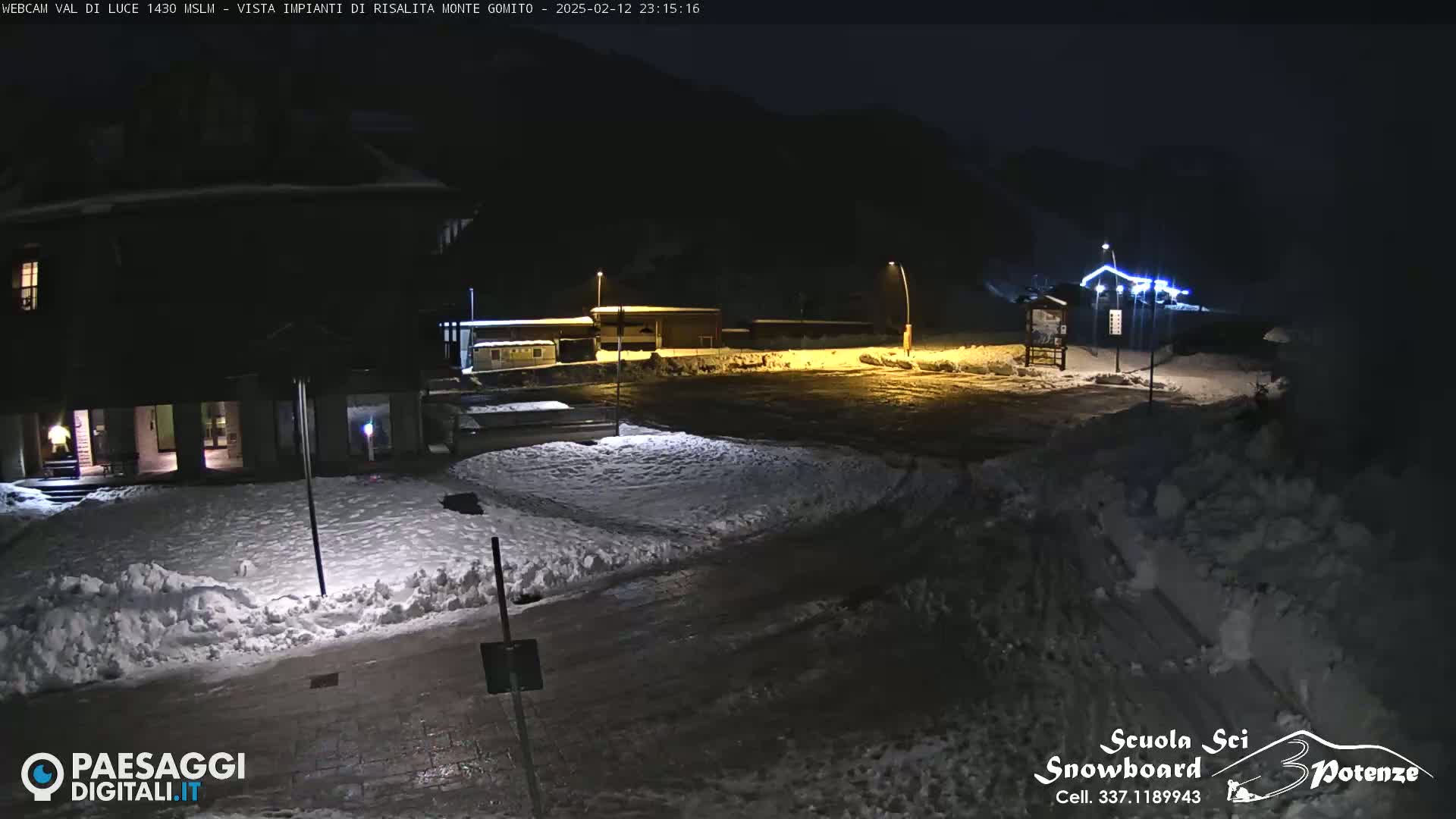 A snow-covered nighttime scene shows a building and a parking lot illuminated by streetlights, with a dark mountainside in the background.