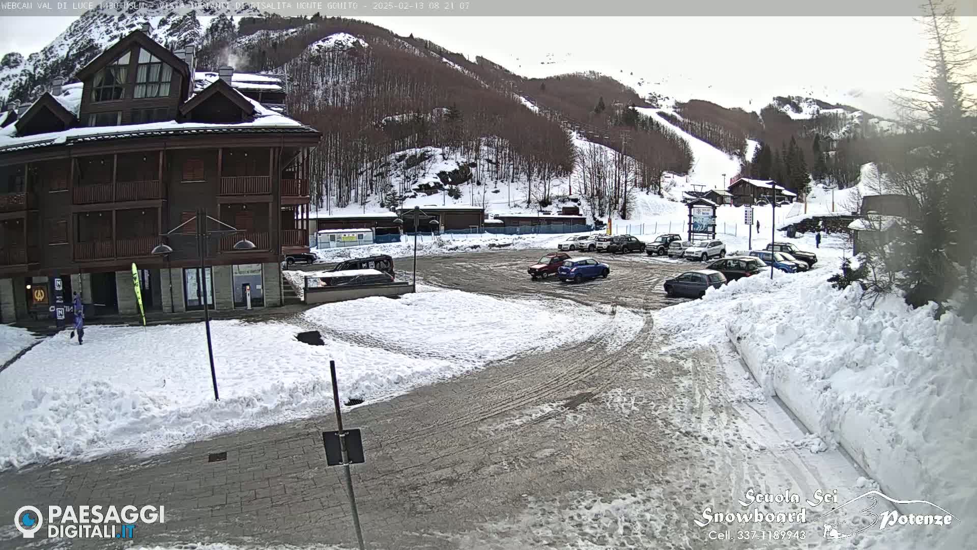 A snow-covered parking lot with several cars is situated in front of a multi-story building, with a snowy mountainside visible in the background.