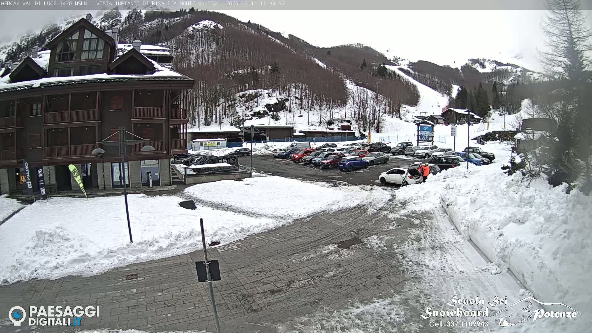 A snow-covered parking lot with cars and a multi-story building is situated in front of a snow-dusted mountain slope.