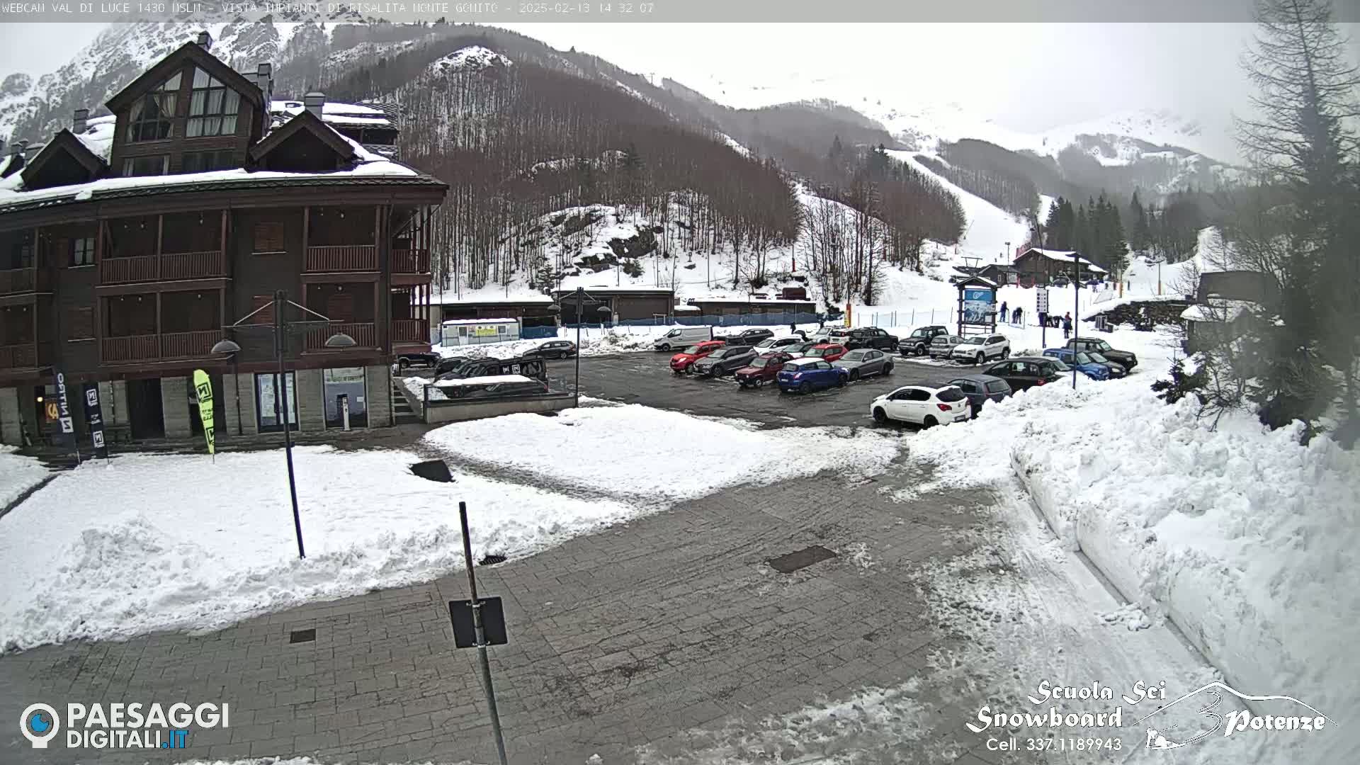 A snow-covered parking lot with cars is in front of a multi-story building, with snow-covered mountains and ski slopes in the background.